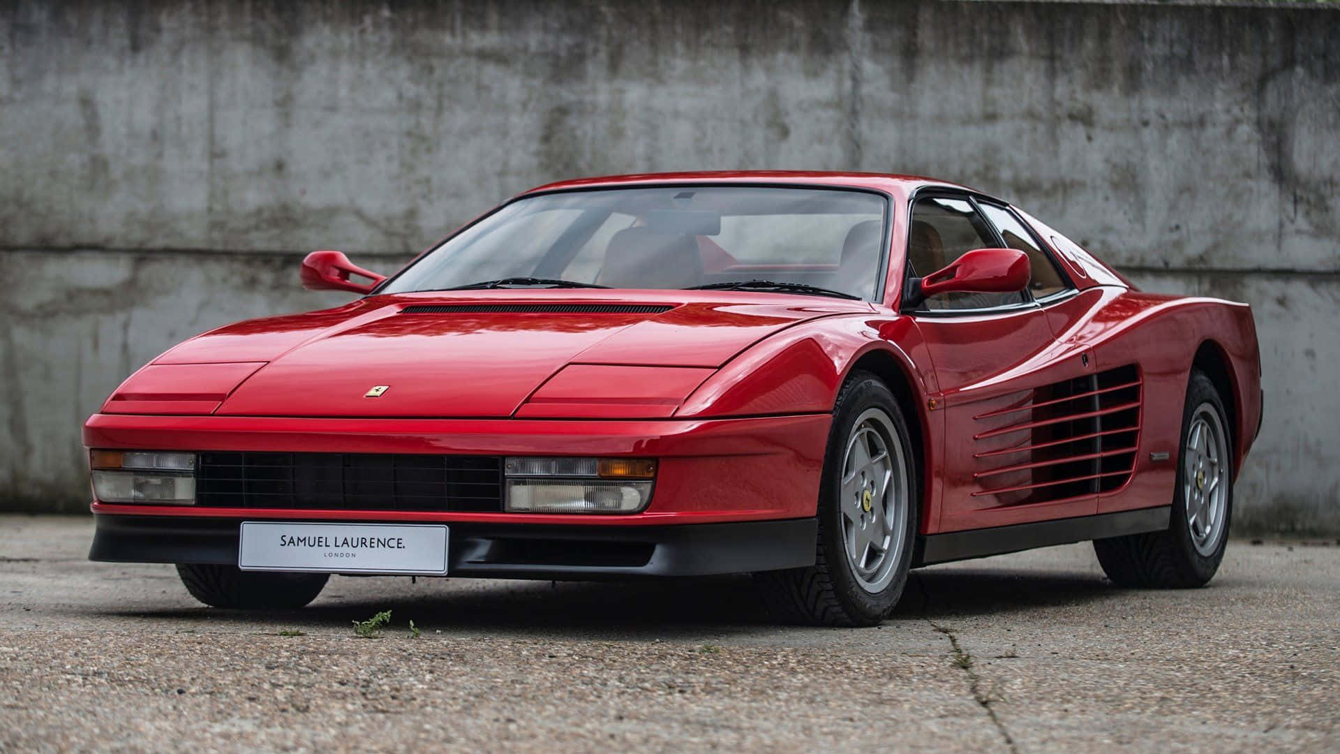 Ferrari Testarossa 桌布 壁紙 — 一輛亮麗的紅色法拉利Testarossa停靠在風化混凝土牆旁的碎石地面上。