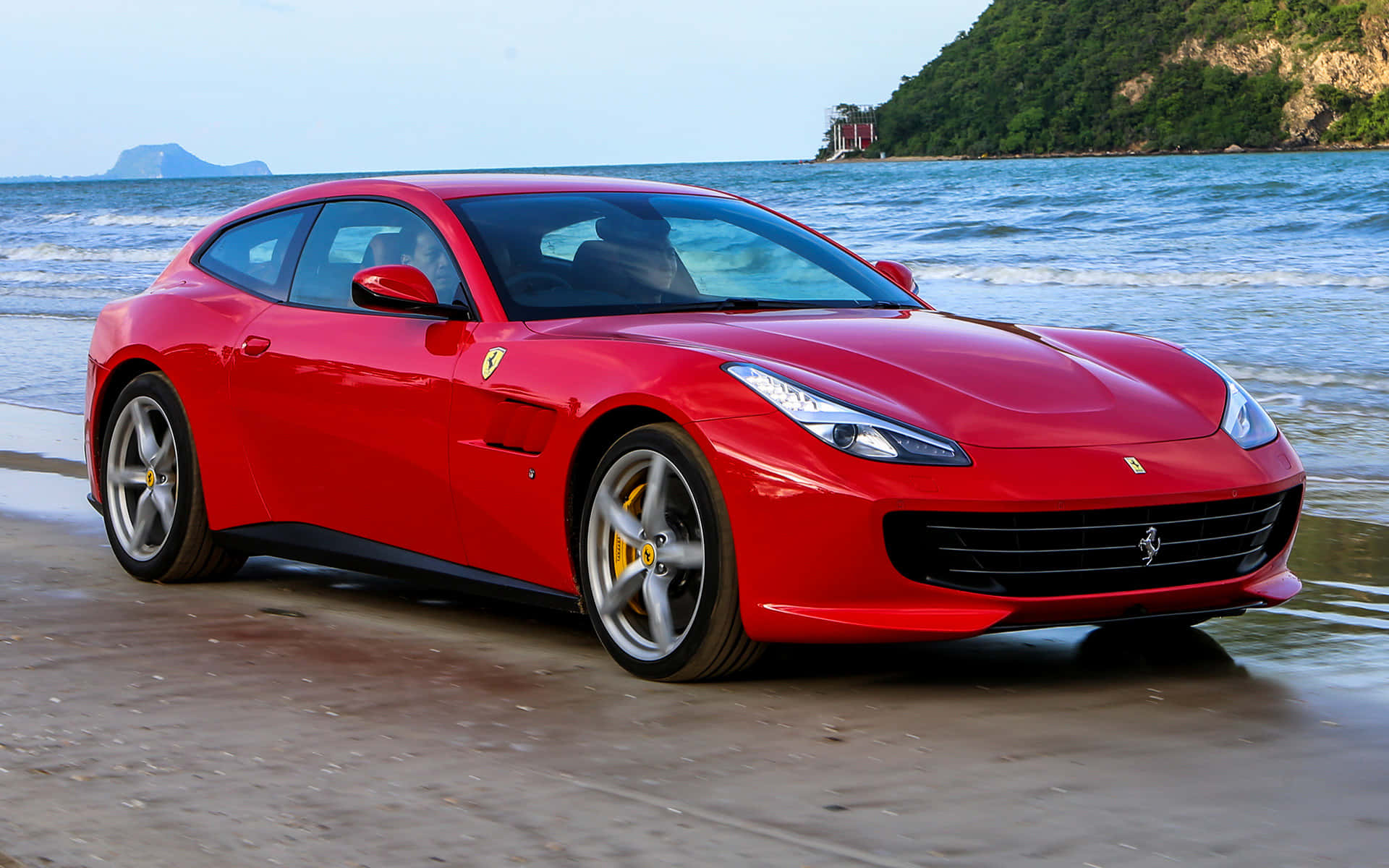 Ferrari Gtc4lusso 桌布 壁紙 — 一輛亮麗的紅色法拉利跑車停泊在濕潤的沙灘上，背景有海浪和綠色山丘。