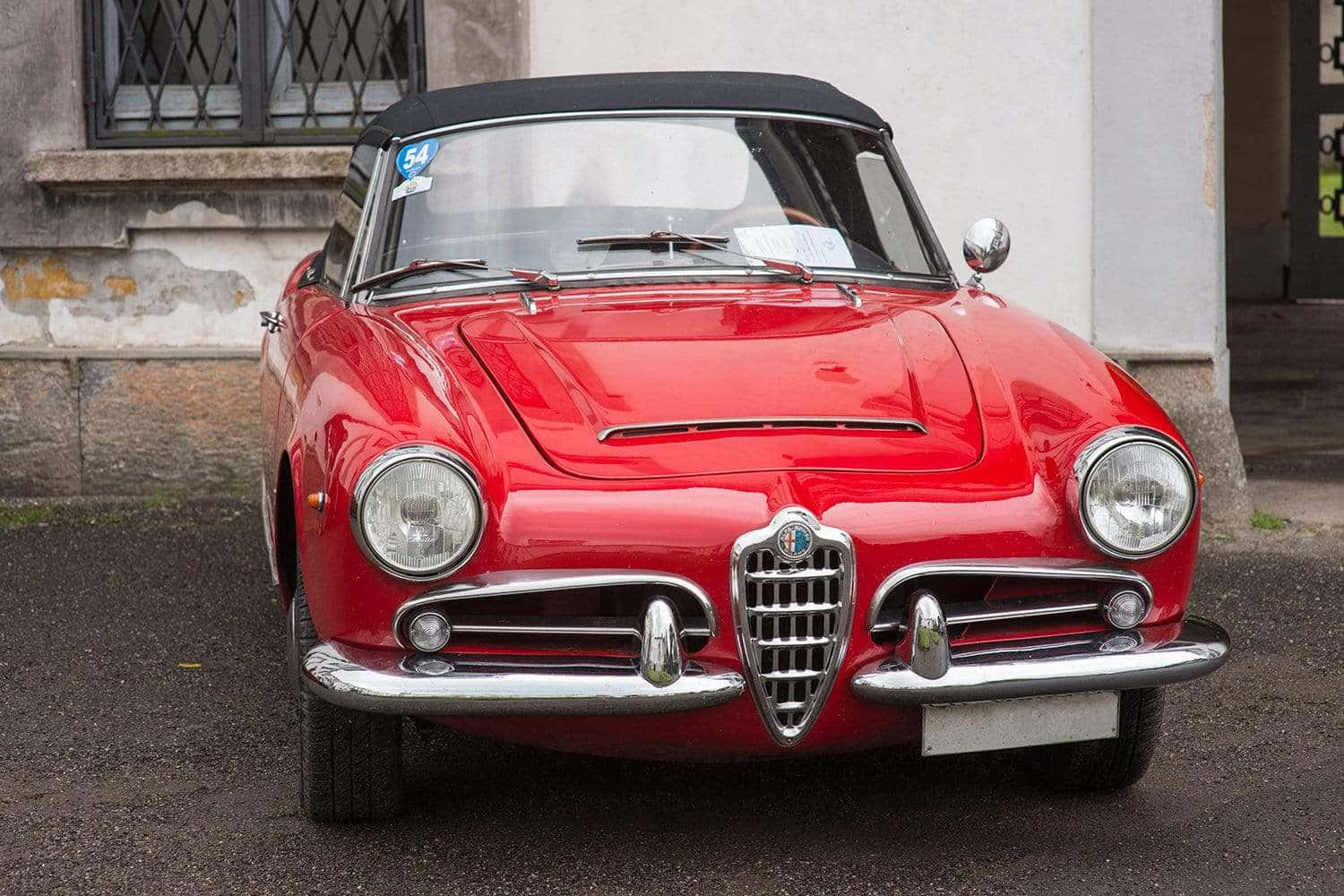 Alfa Romeo Spider 桌布 壁紙 — 一輛亮麗的紅色經典愛快羅密歐Spider敞篷車停放在潮濕的街道上，旁邊是一棟風化的建築物。