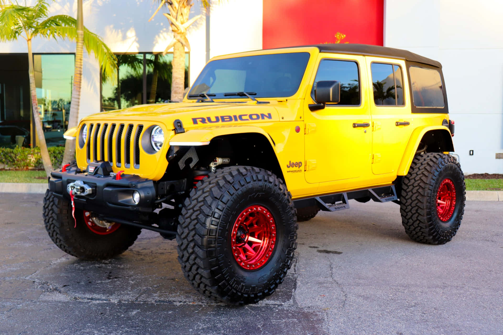 Jeep Wrangler 桌布 壁紙 — 一輛亮黃色Jeep Rubicon配備紅色輪圈和超大輪胎，停在棕櫚樹和現代建築附近的瀝青路面上。