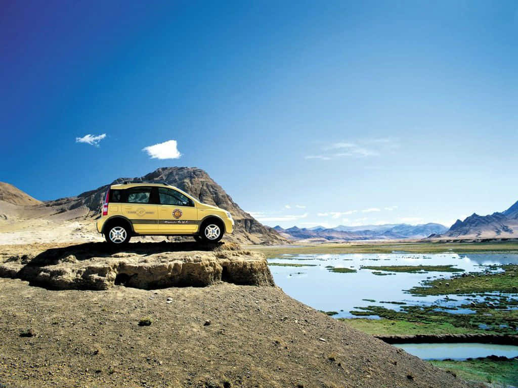 Fiat Panda 桌布 壁紙 — 一輛亮黃色SUV停靠在岩石露頭上，俯瞰著寧靜的湖泊，遠處山巒在清澈的藍天下。