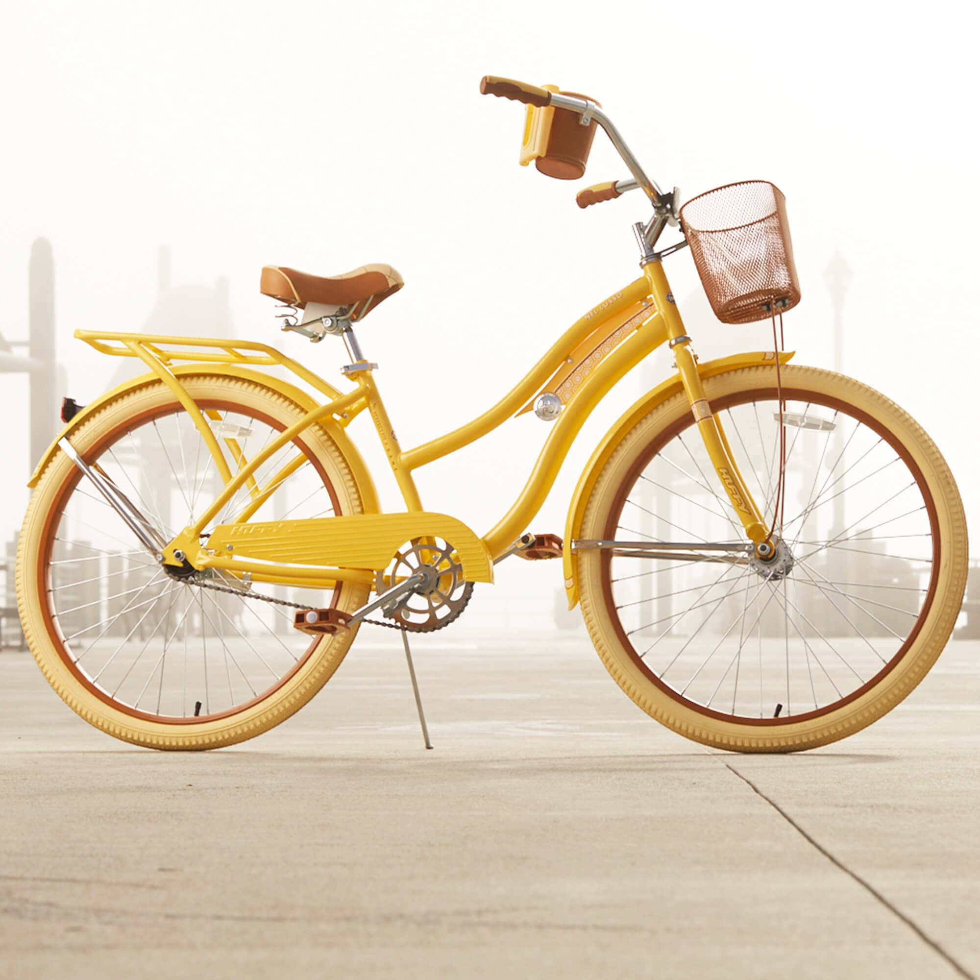 Beach Cruiser 桌布 壁紙 — 一輛亮黃色復古自行車，配有棕色籃子和座椅，立在混凝土表面上，背景是朦朧的城市景觀。