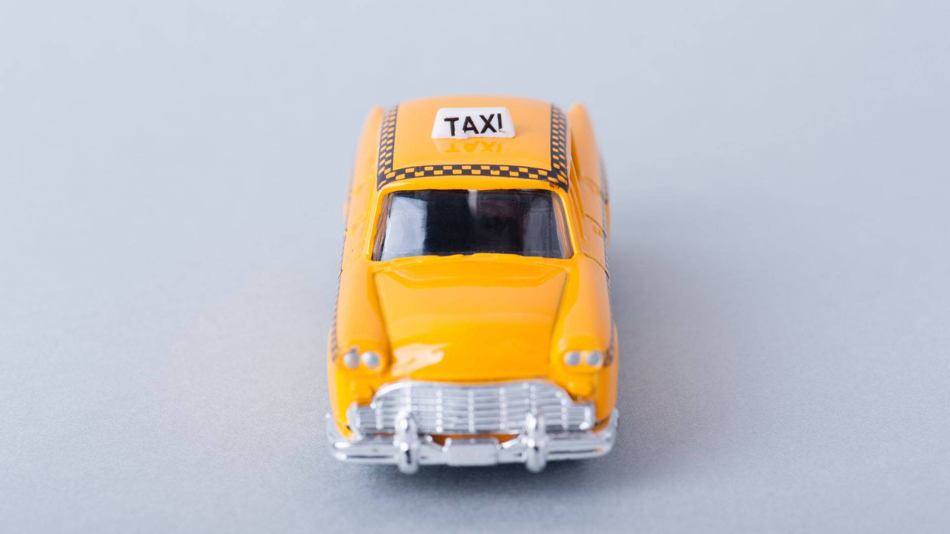 taxi 桌布 壁紙 — 一輛亮黃色復古計程車玩具車，帶有黑色棋盤格車頂和銀色格柵，居中放置在純灰色表面上。
