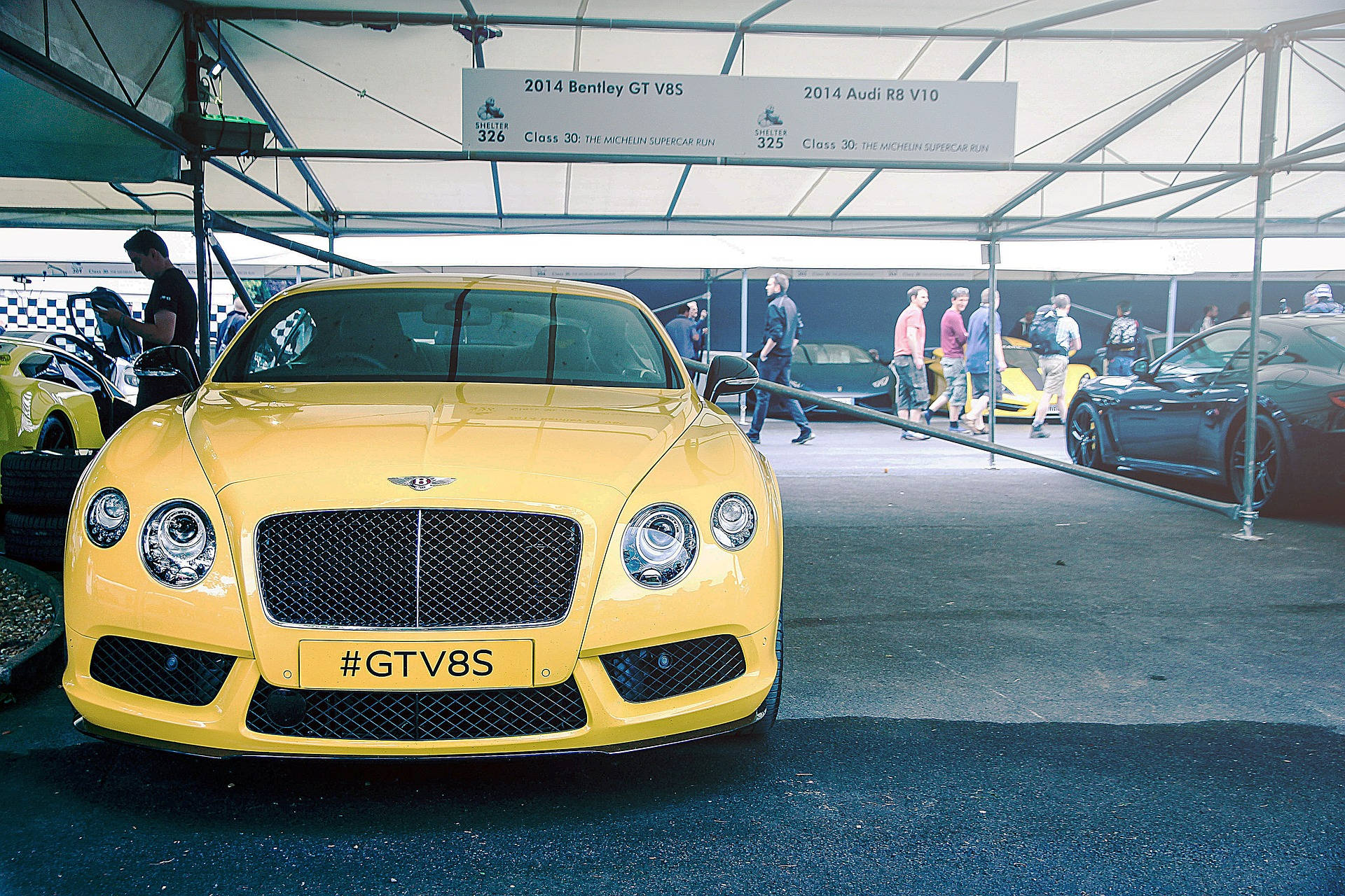 bentley HD 桌布 壁紙 — 一輛亮黃色的2014年賓利GT V8S在汽車集會的帳篷下閃閃發光，附近有觀眾和其他豪華車輛。
