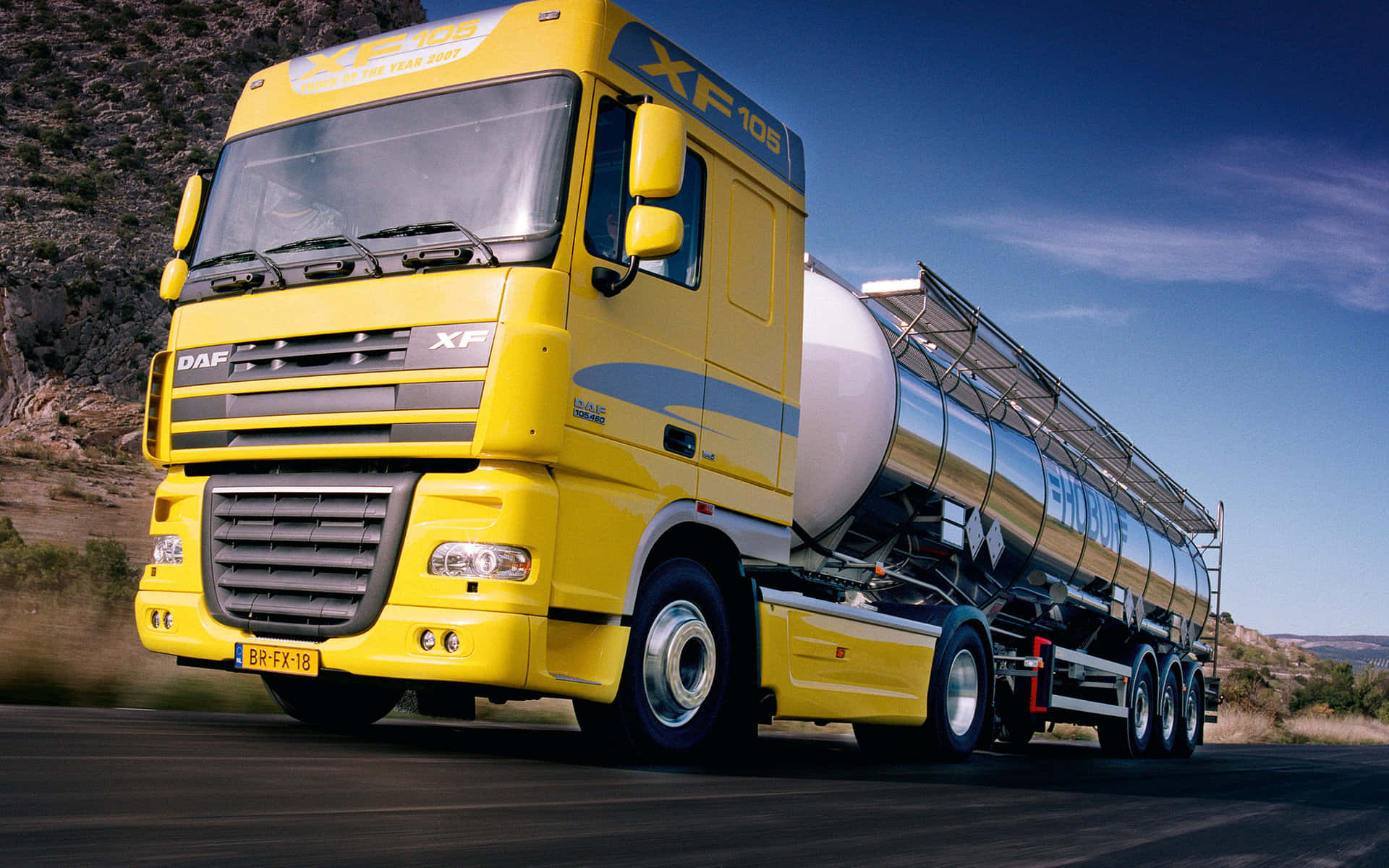 Semi Truck 桌布 壁紙 — 一輛亮黃色的DAF XF105油罐車，帶有銀色油罐，在晴朗藍天下行駛在乾燥丘陵的道路上。