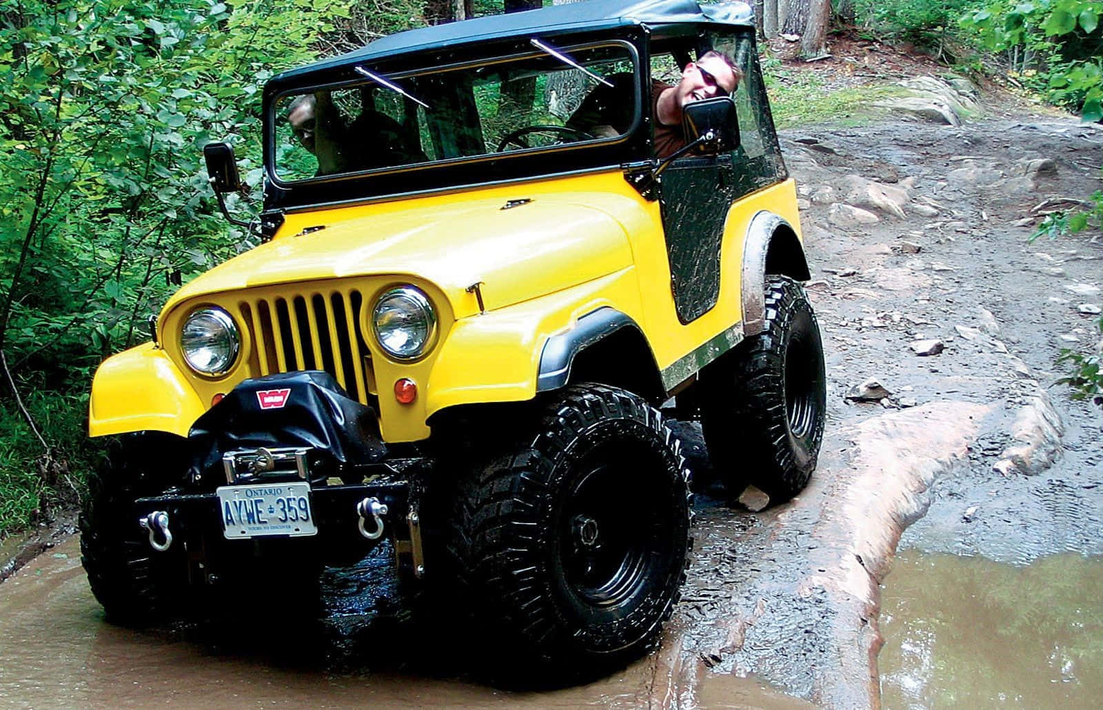 Jeep Cj 桌布 壁紙 — 一輛亮黃色的Jeep Wrangler在泥濘的森林小徑上行駛，司機面帶微笑享受越野冒險。