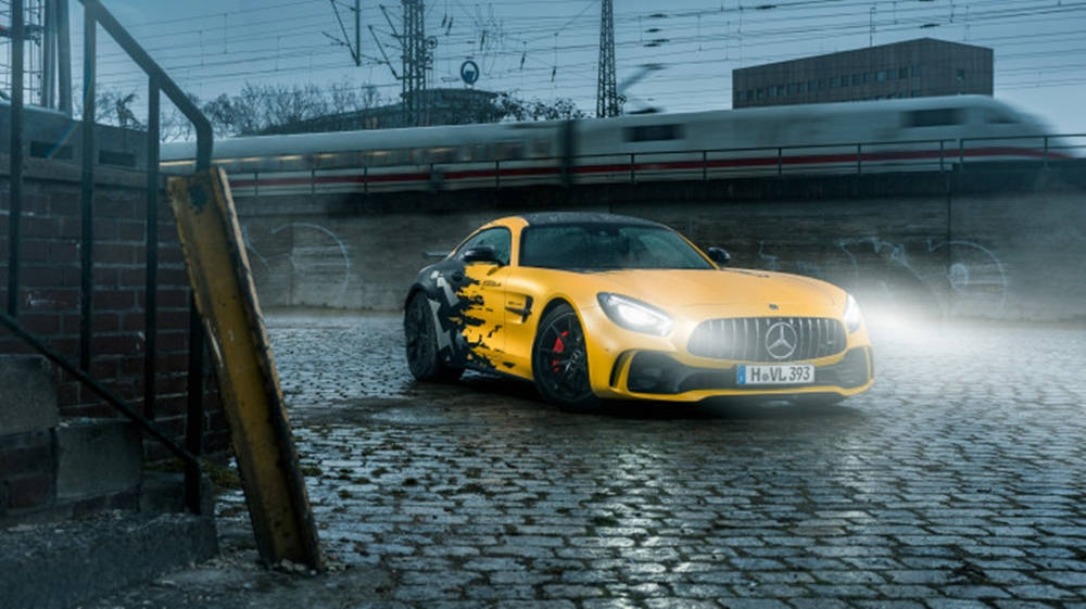 Gtr 桌布 壁紙 — 一輛亮黃色的Mercedes-AMG GT跑車帶有黑色火焰貼紙，停在濕漉漉的鵝卵石上，一列高速列車模糊地從上方駛過。