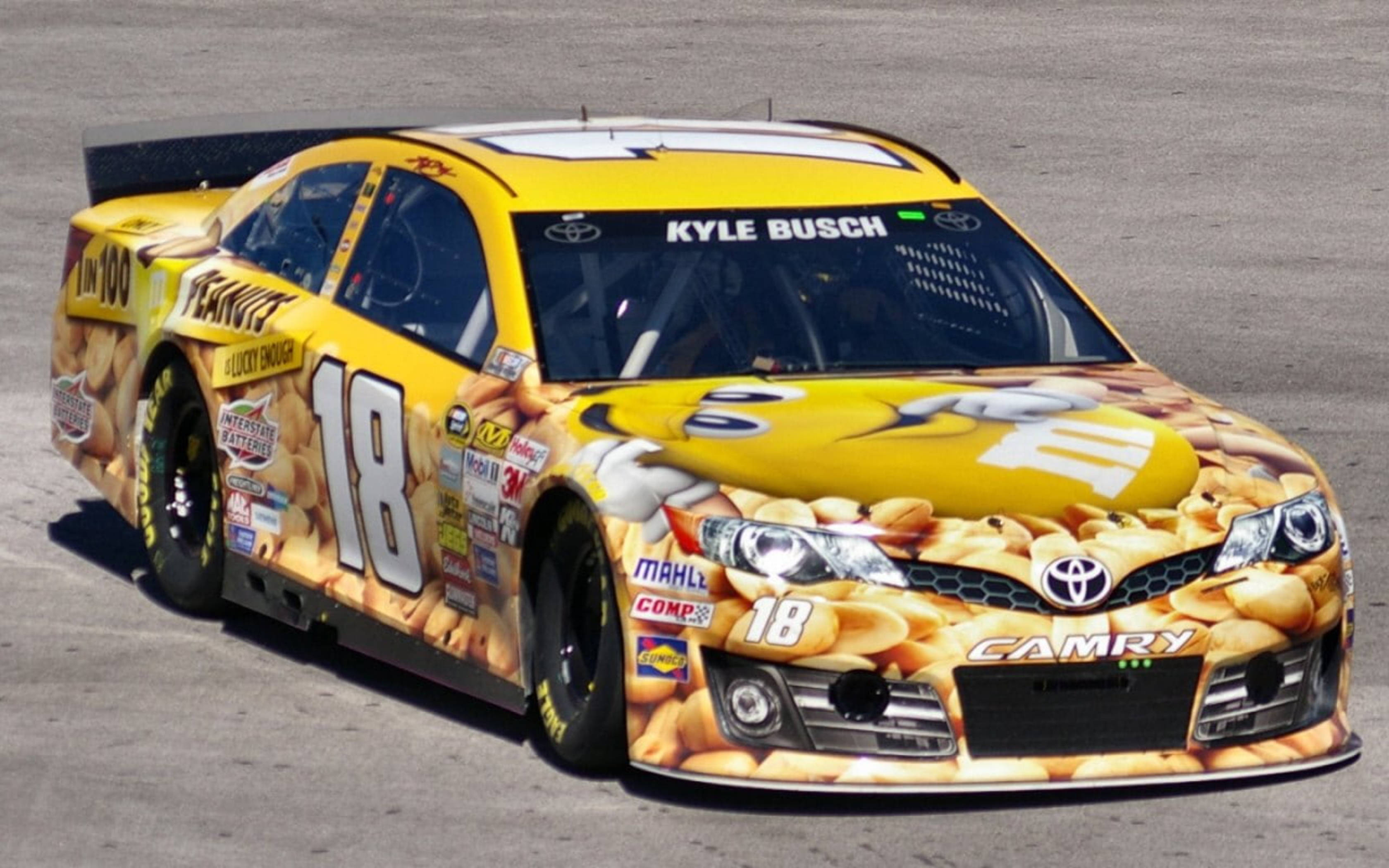 Racing 桌布 壁紙 — 一輛亮黃色的NASCAR豐田Camry賽車，覆蓋著花生醬和Reese's花生醬杯圖案，停在灰色瀝青賽道上。
