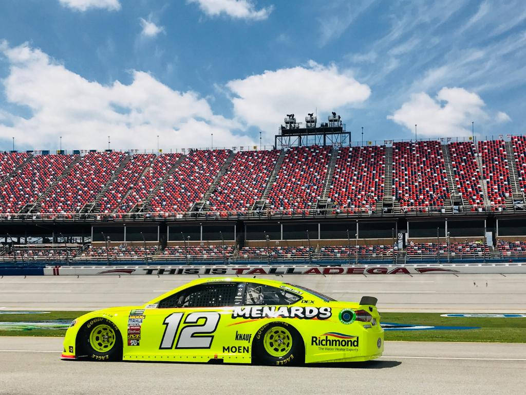 Ryan Blaney 桌布 壁紙 — 一輛亮黃色的NASCAR賽車（12號）在藍天白雲和擠滿紅色座位的體育館下，在賽道上飛馳。