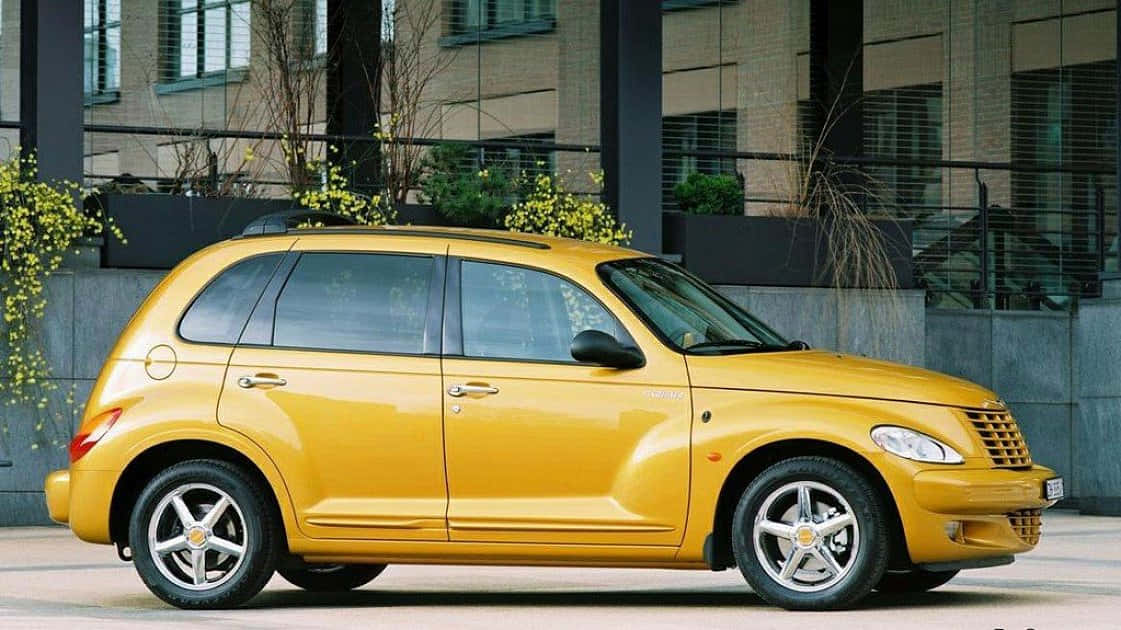 Chrysler Pt Cruiser 桌布 壁紙 — 一輛亮黃色的PT Cruiser停放在現代玻璃建築旁的城市人行道上，旁邊有開花的灌木叢。