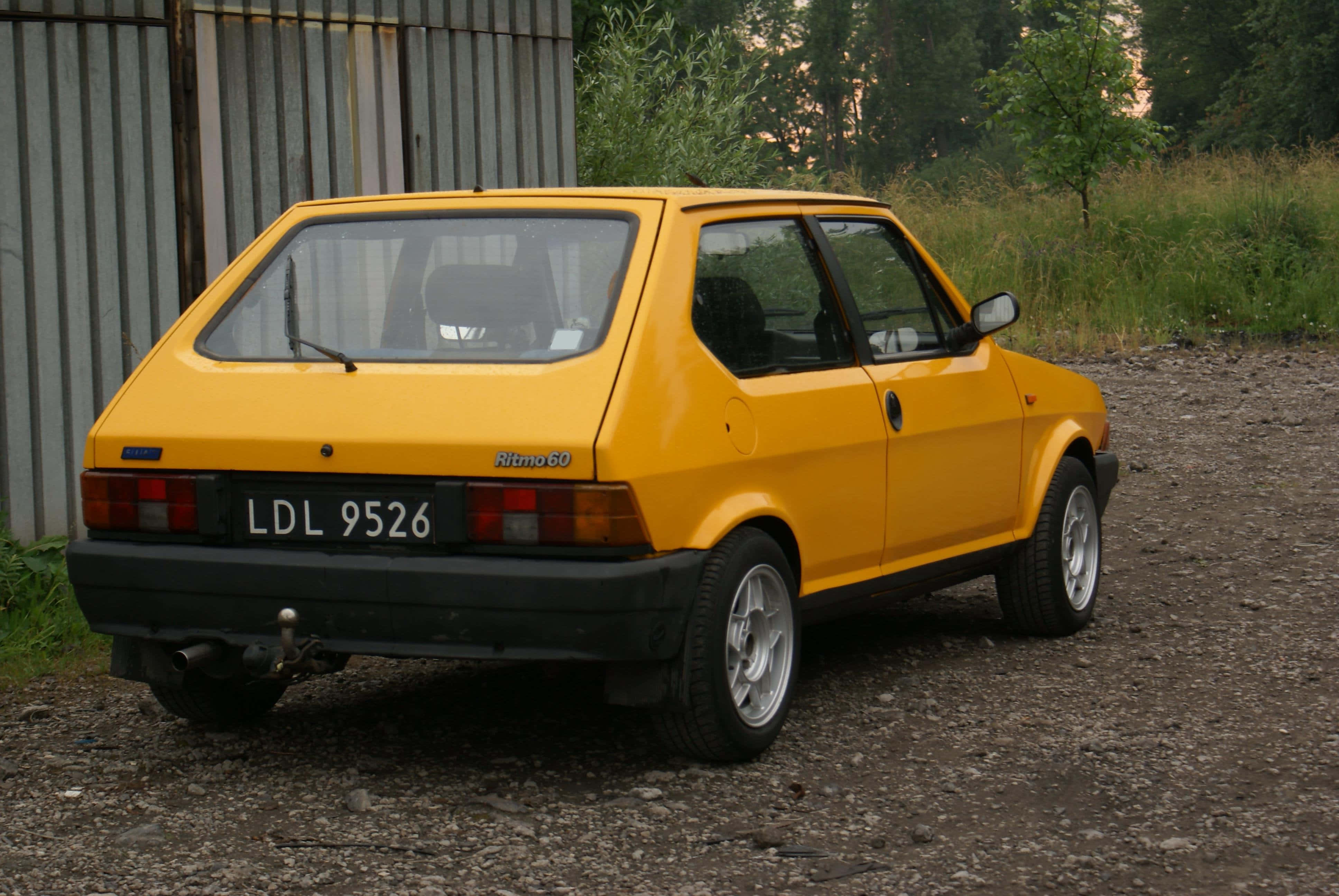 Fiat Ritmo 桌布 壁紙 — 一輛亮黃色的快意 Ritmo 60 掀背車在黃昏時停泊在碎石地上，旁邊是波紋金屬圍欄。