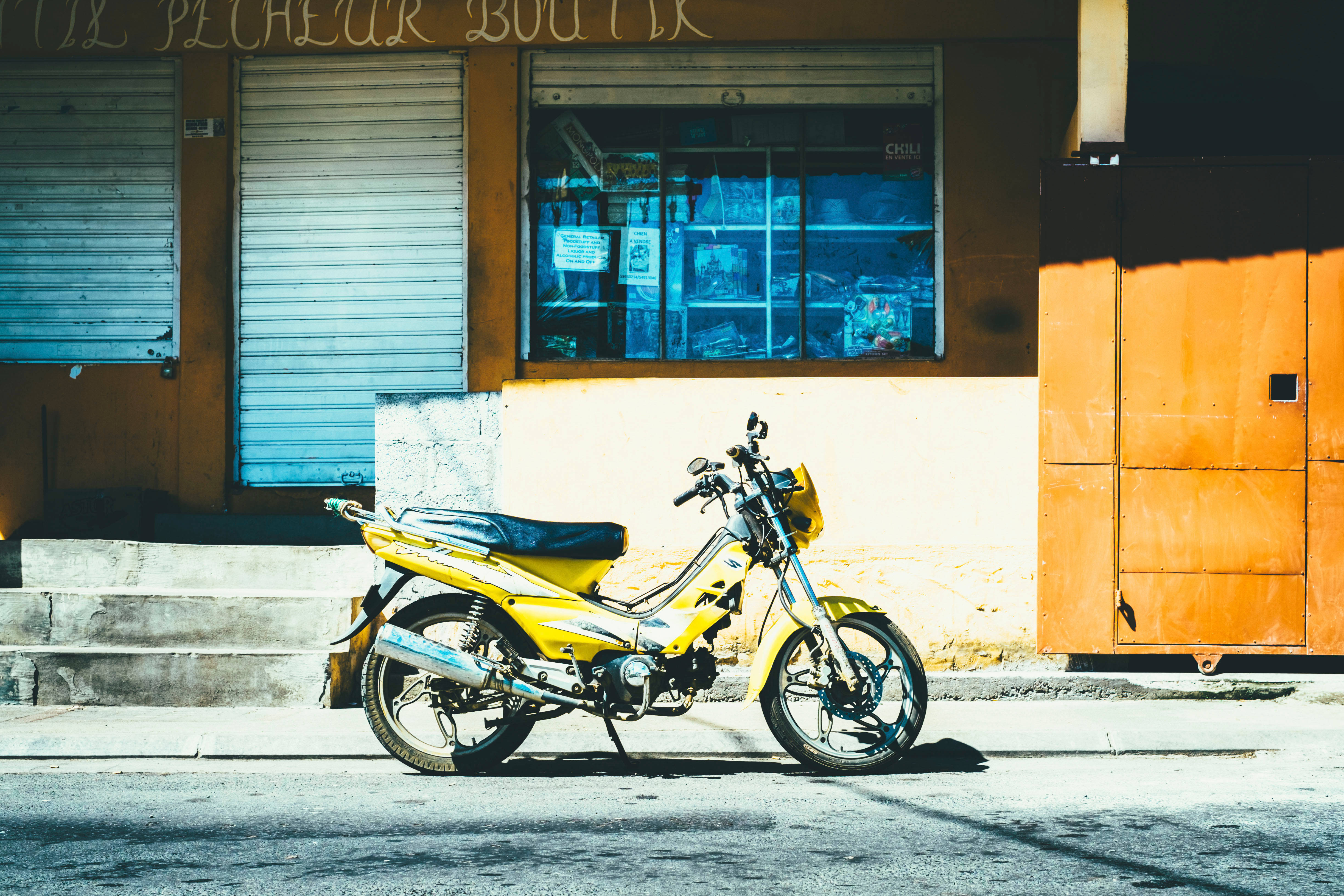 機車 桌布 壁紙 — 一輛亮黃色的摩托車停放在陽光明媚的街道上，旁邊是一家老舊的商店，百葉窗關閉，窗戶帶有藍色色調。