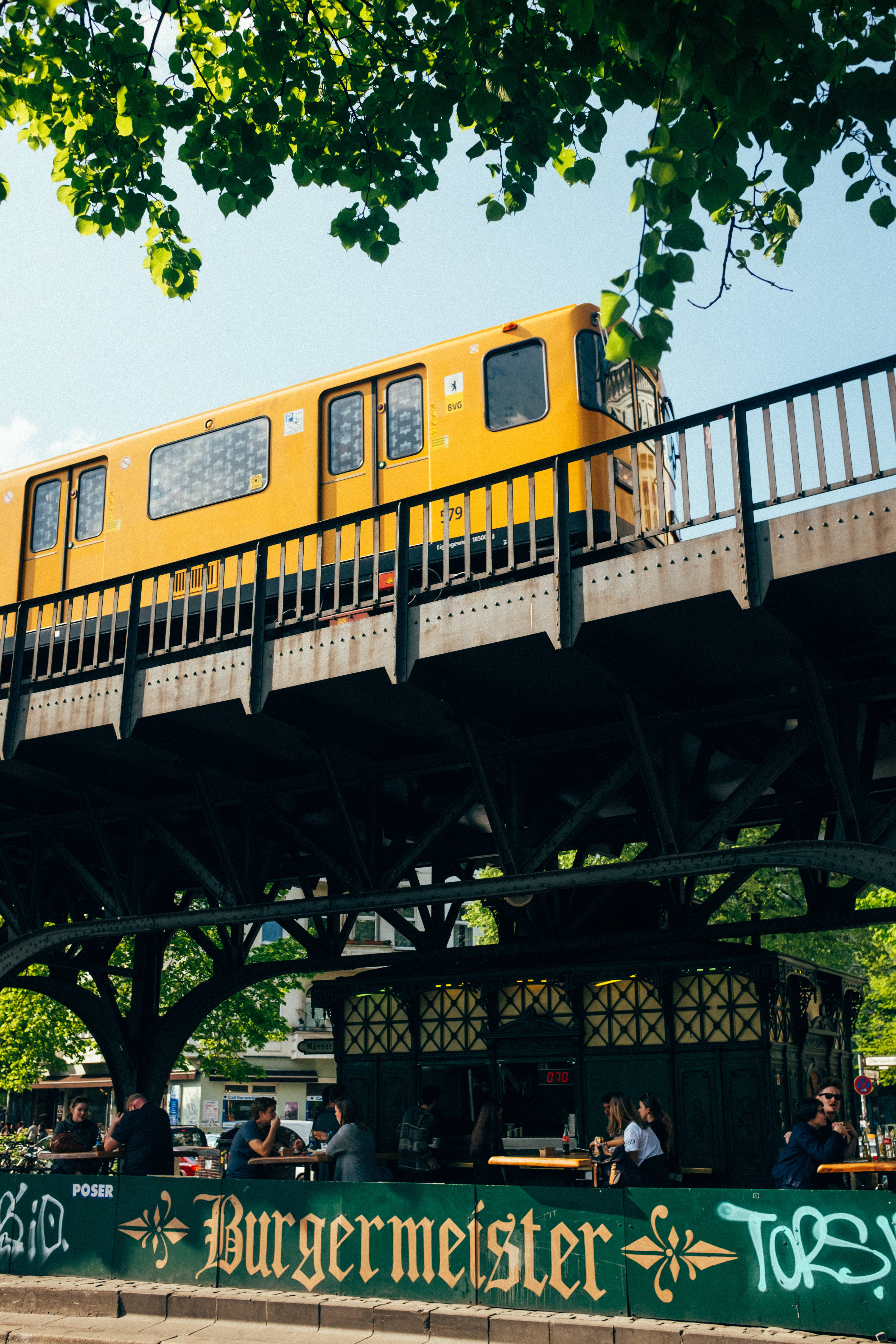 Berlin 桌布 壁紙 — 一輛亮黃色的火車在高架軌道上滑行，下方是熱鬧的戶外咖啡館，被茂密的綠樹遮蔭。