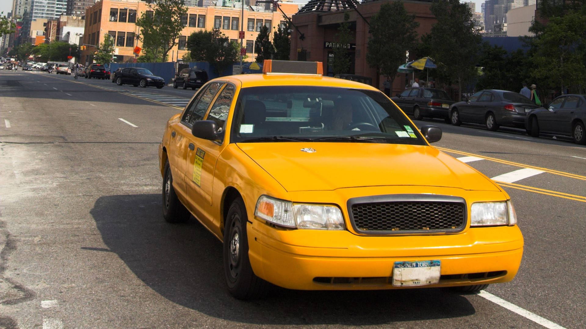 Road 桌布 壁紙 — 一輛亮黃色的計程車在陽光普照的市區街道上行駛，街道兩旁有建築物和樹木。