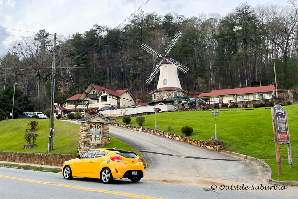 風景 桌布 壁紙 — 一輛亮黃色的車駛過路邊的汽車旅館，旅館在草地山丘上，有一座高風車，天空多雲。