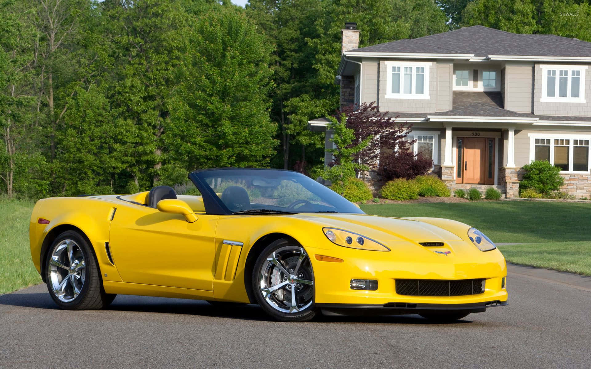 Chevrolet Corvette Grand Sport 桌布 壁紙 — 一輛亮黃色的雪佛蘭克爾維特敞篷車停放在郊區房屋前的車道上，周圍有茂密的綠樹。