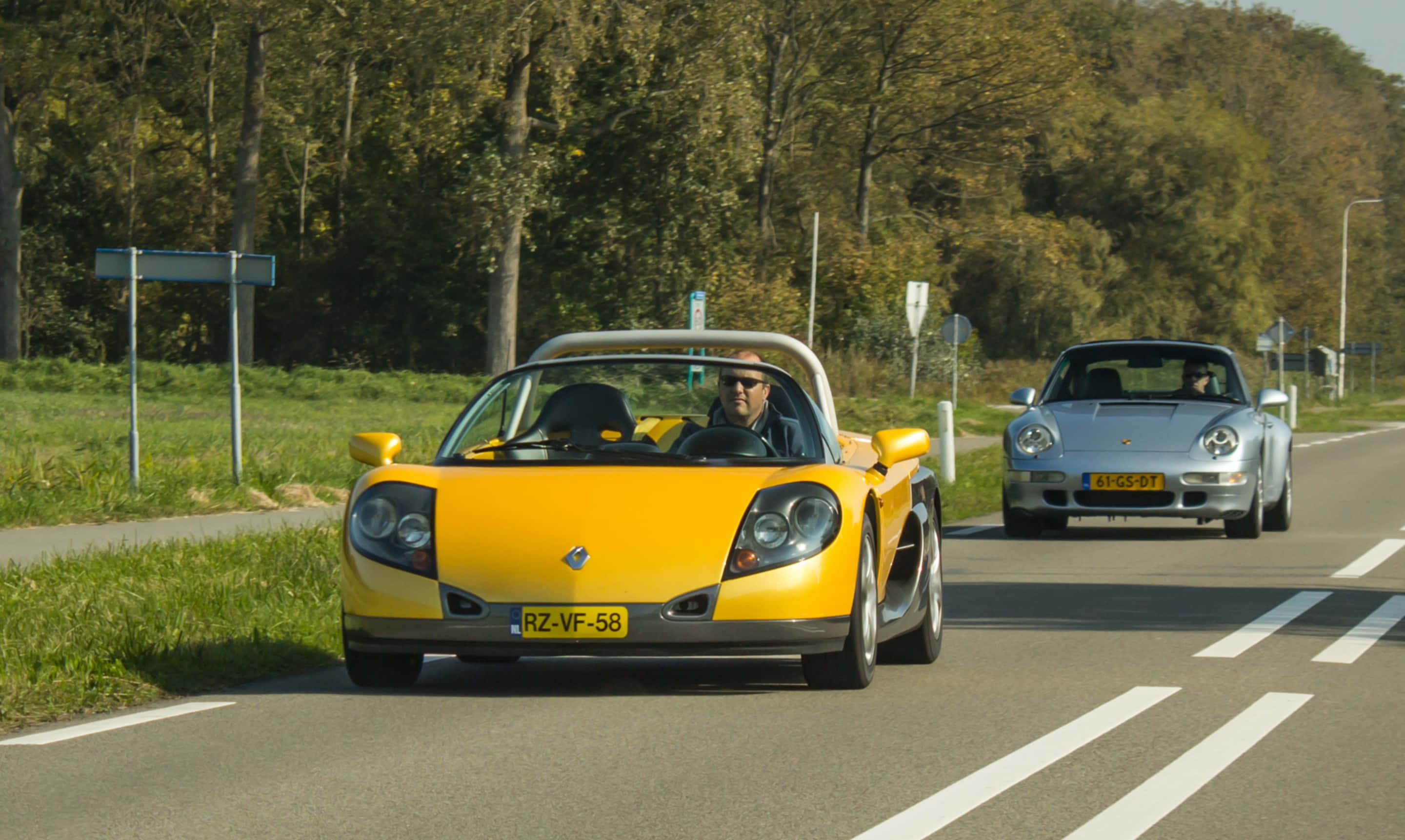 Renault Sport Spider 桌布 壁紙 — 一輛亮黃色的雷諾Spider和銀色的保時捷911 Carrera在晴朗的天空下並排行駛在綠樹成蔭的道路上。