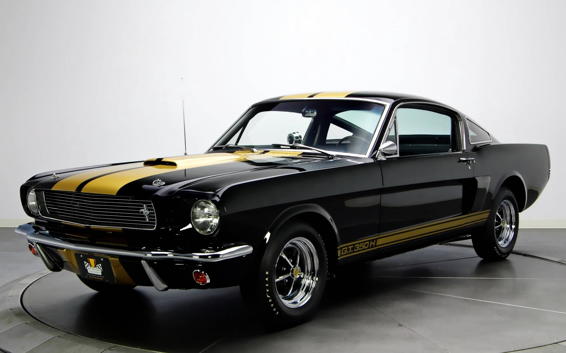 shelby iPhone 桌布 壁紙 — 一輛亮黑色1967年福特Mustang GT350H，帶有醒目的金色賽車條紋，在攝影棚燈光下於圓形轉盤上閃閃發光。