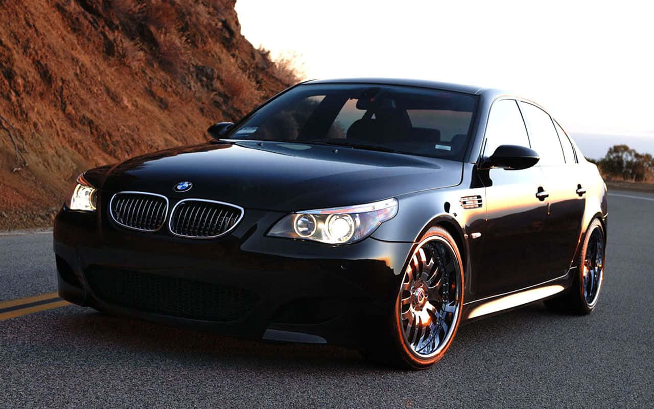 Bmw M5 桌布 壁紙 — 一輛亮黑色BMW M5轎車配備定制輪圈，在黃金時段沿著蜿蜒的道路閃耀，背景是崎嶇的懸崖。