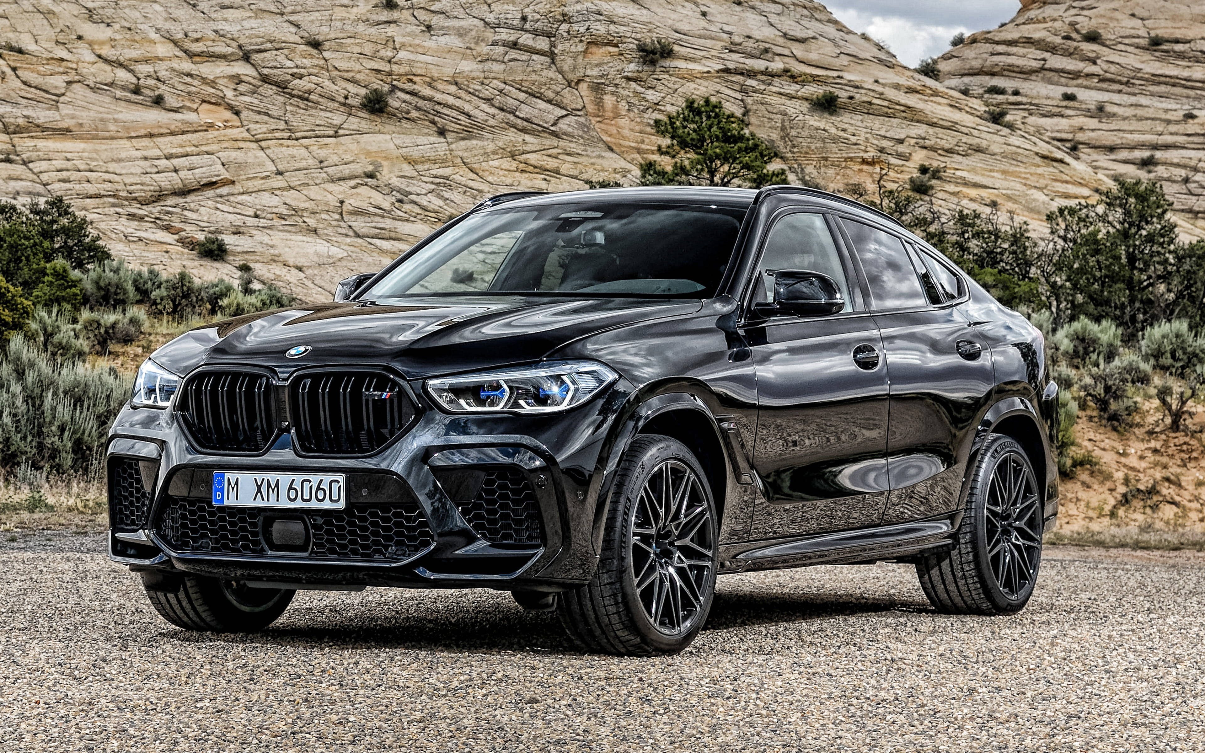 m 桌布 壁紙 — 一輛亮黑色BMW X6 M停在礫石上，背景有堅固的岩層和稀疏的沙漠植被。