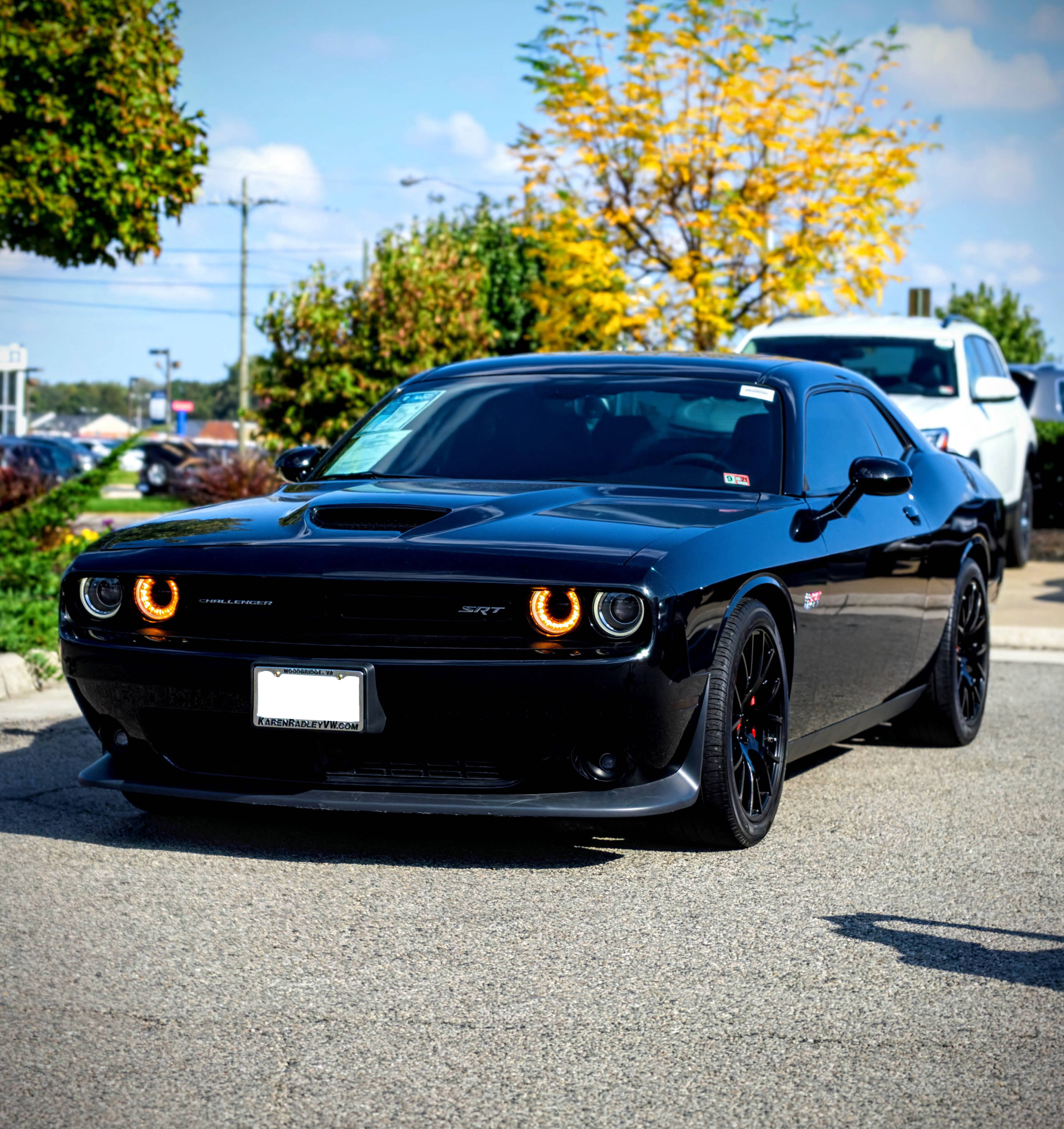 mustang HD 桌布 壁紙 — 一輛亮黑色Dodge Challenger SRT，帶有發光的橙色光環頭燈，停在柏油路面上，背景是明亮的藍天和秋樹。