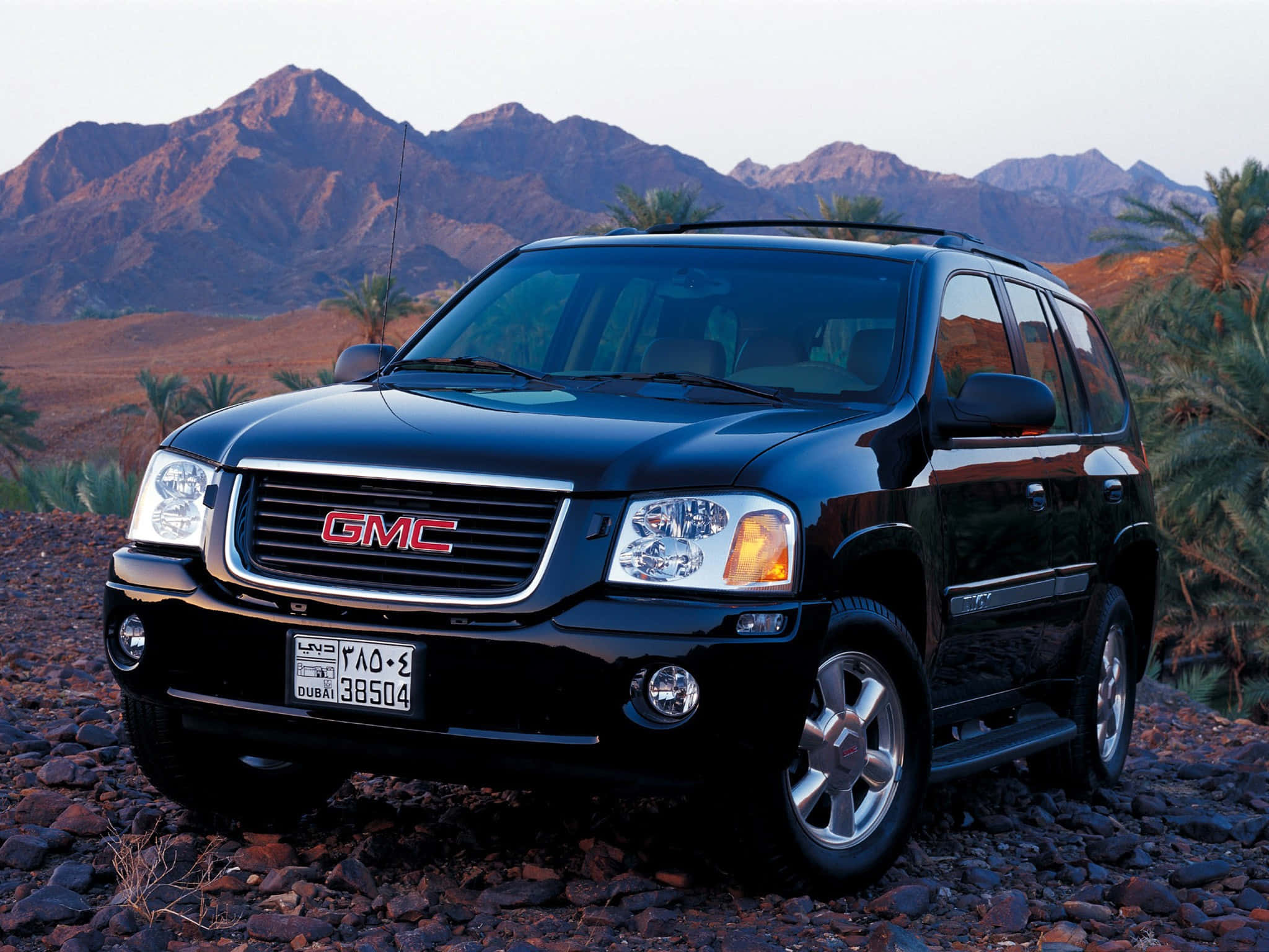 Gmc Envoy 桌布 壁紙 — 一輛亮黑色GMC Envoy SUV停泊在岩石沙漠地形上，背景有棕櫚樹和崎嶇山脈，正值日落時分。