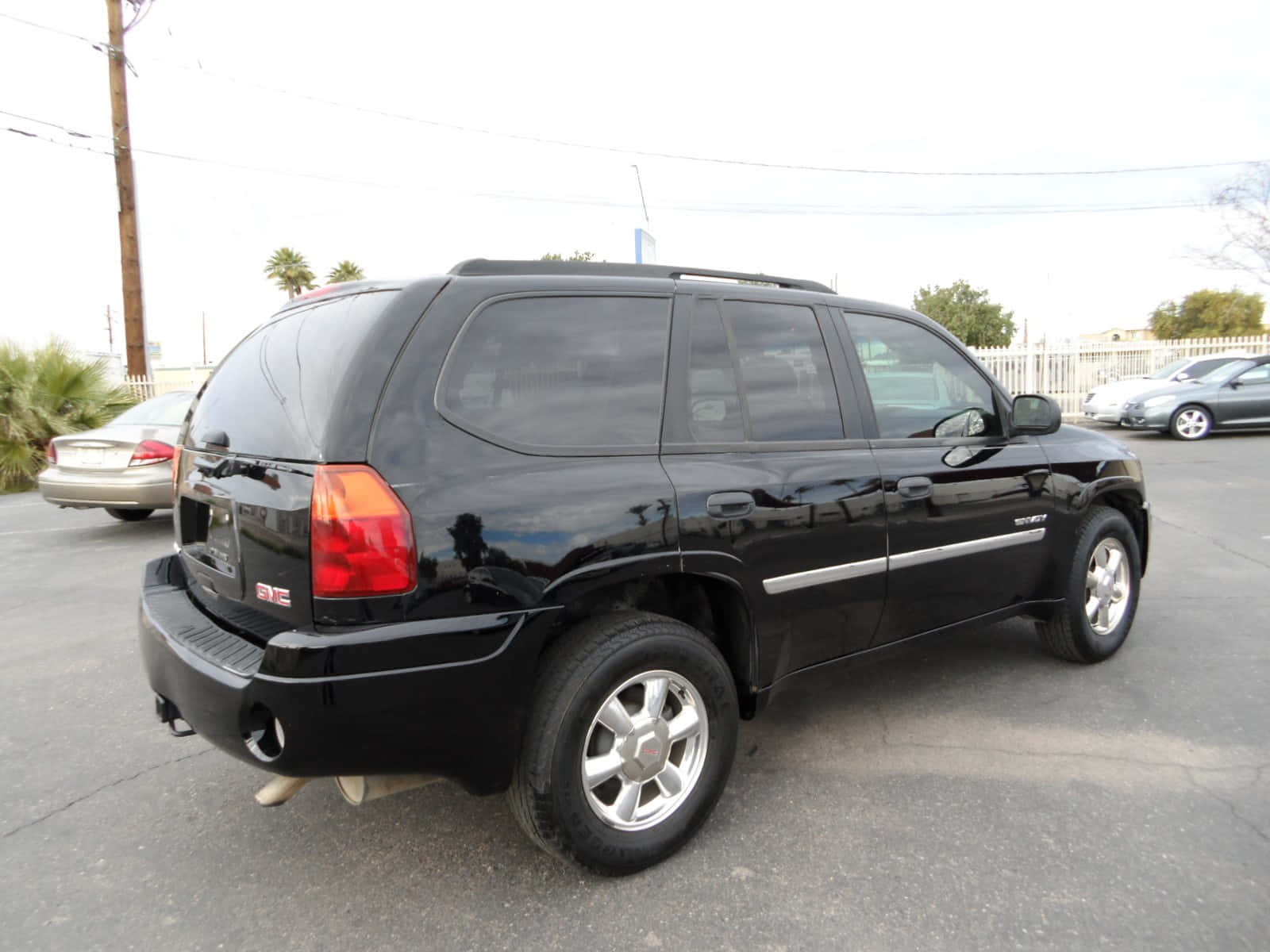 Gmc Envoy 桌布 壁紙 — 一輛亮黑色GMC Envoy SUV停放在停車場，背景有棕櫚樹和其他車輛，天空多雲。