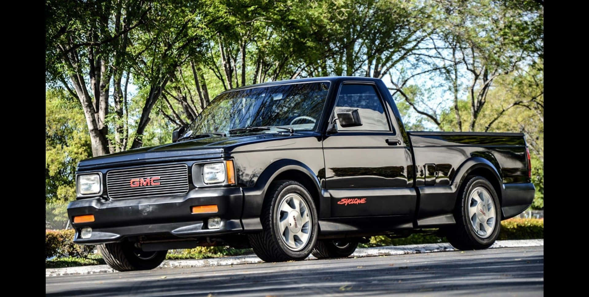 Gmc Syclone 桌布 壁紙 — 一輛亮黑色GMC Syclone皮卡車停在鋪砌的道路上，在枝葉繁茂的綠樹下，天氣晴朗。