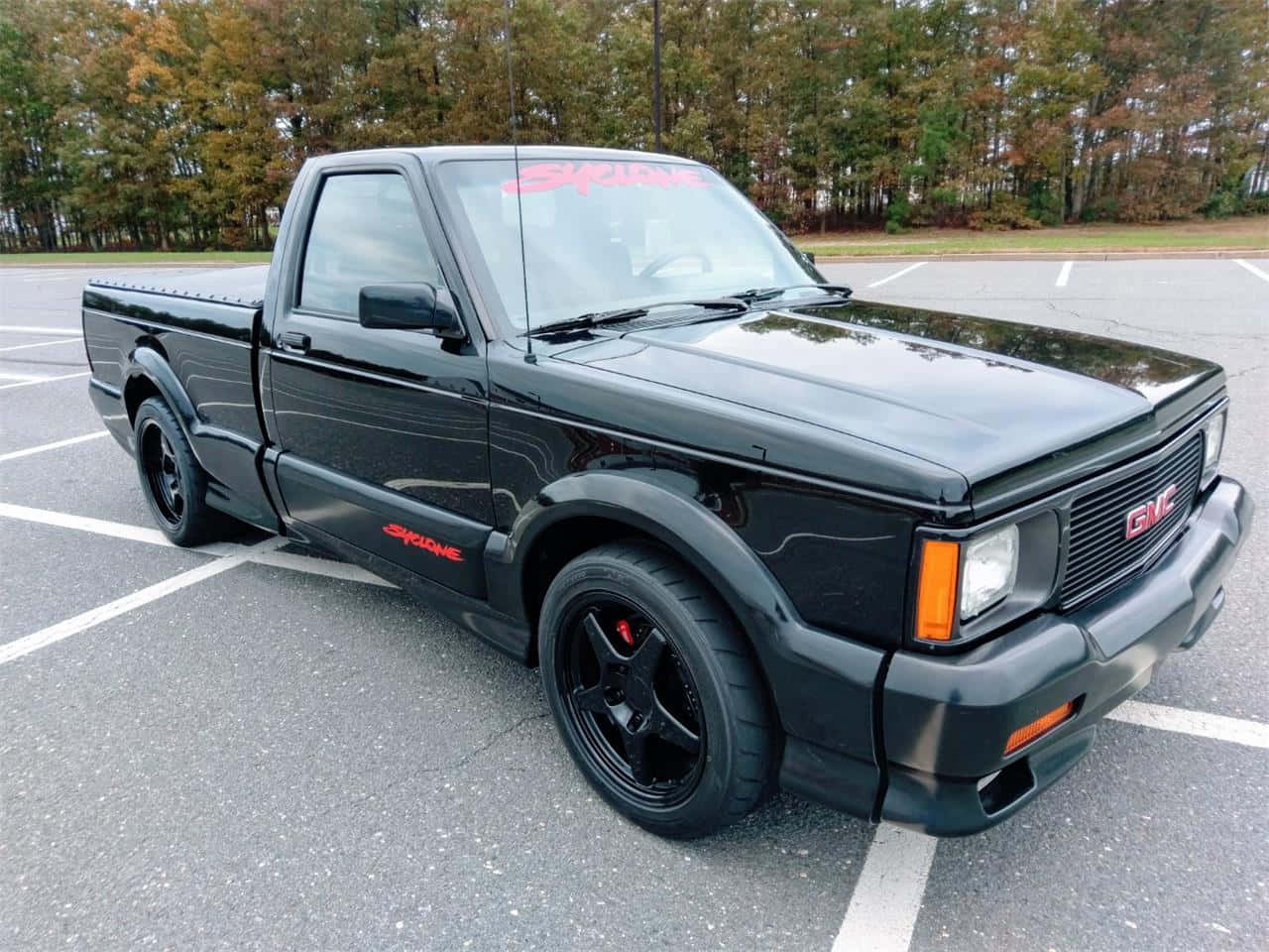 黑色GMC Syclone皮卡車帶有紅色貼紙，停在一個被秋樹環繞的停車場。 — Toyota Supra Mk4 壁紙