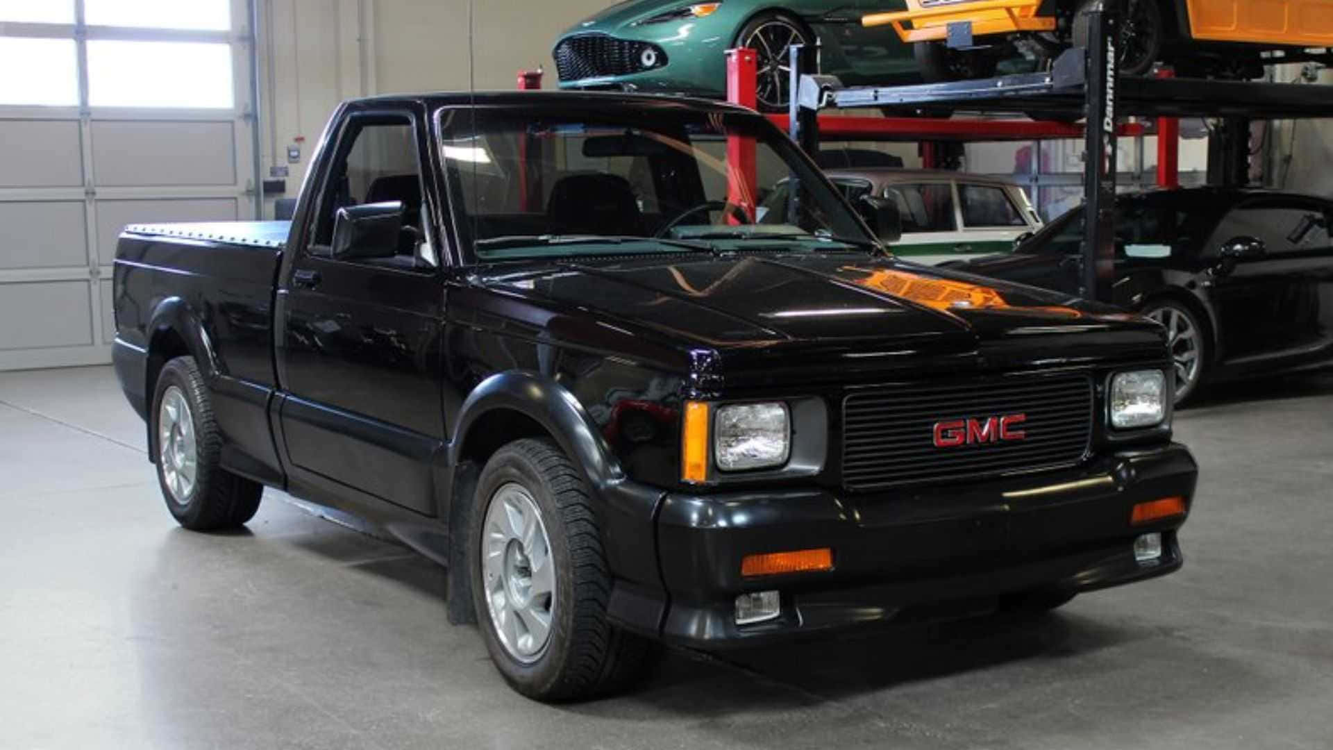 Gmc Syclone 桌布 壁紙 — 一輛亮黑色GMC皮卡車停放在光線充足的車庫中，背景有其他車輛在升降機上。