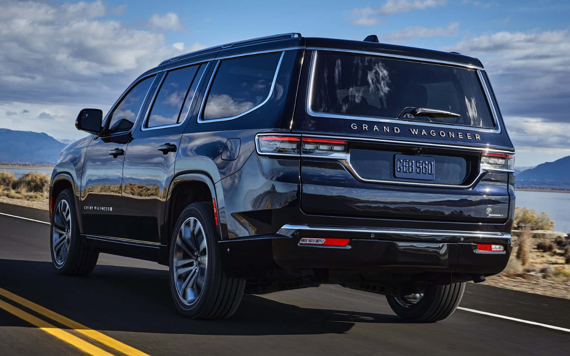 Jeep Wagoneer 桌布 壁紙 — 一輛亮黑色Grand Wagoneer SUV在沙漠公路上巡航，背景有山脈和水域，天空部分多雲。