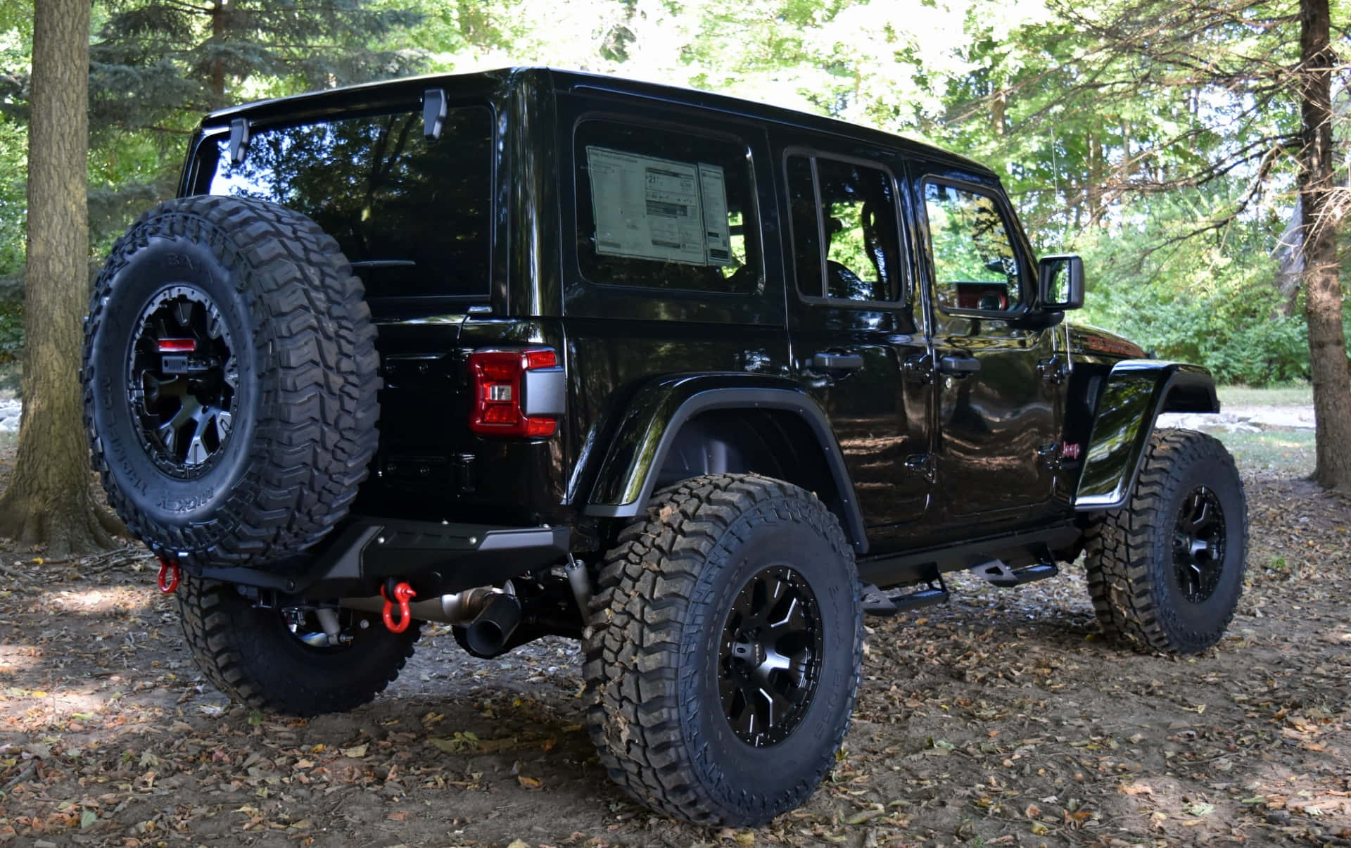 Jeep Wrangler 桌布 壁紙 — 一輛亮黑色Jeep Wrangler配備超大輪胎，停放在樹葉茂密的森林地面上，陽光斑駁灑落。