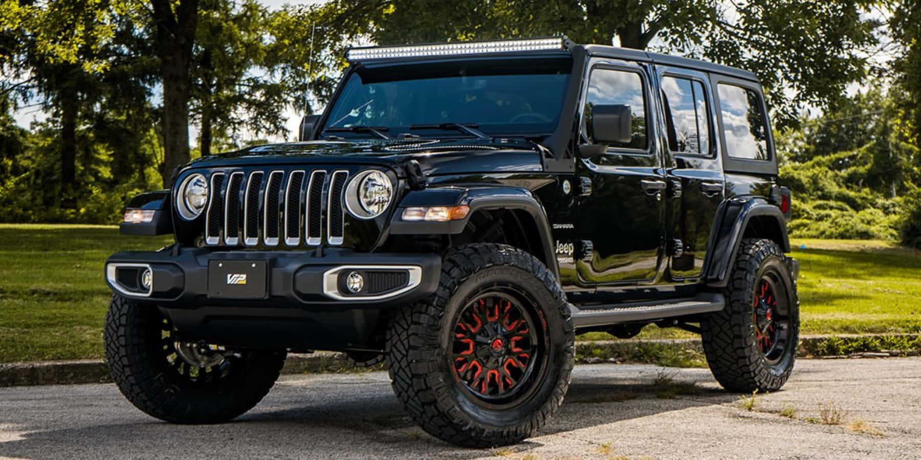 黑色吉普車 桌布 壁紙 — 一輛亮黑色Jeep Wrangler配紅色輪圈，在陽光明媚的日子停放在鋪滿樹蔭的柏油路上。