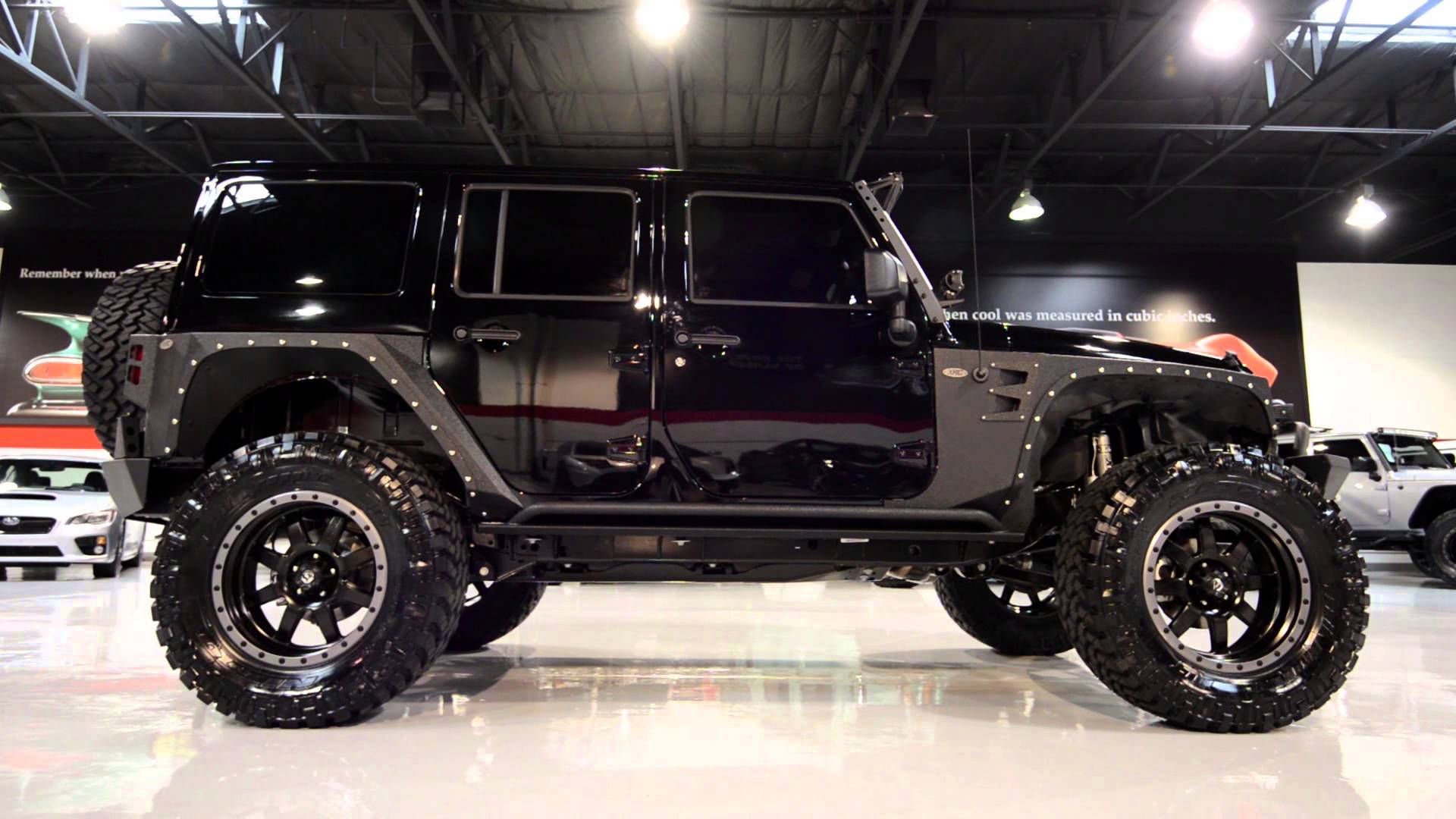 Wrangler 桌布 壁紙 — 一輛亮黑色升高底盤的Jeep Wrangler，配備超大輪胎，在明亮的頂燈下展示於展廳地板上。