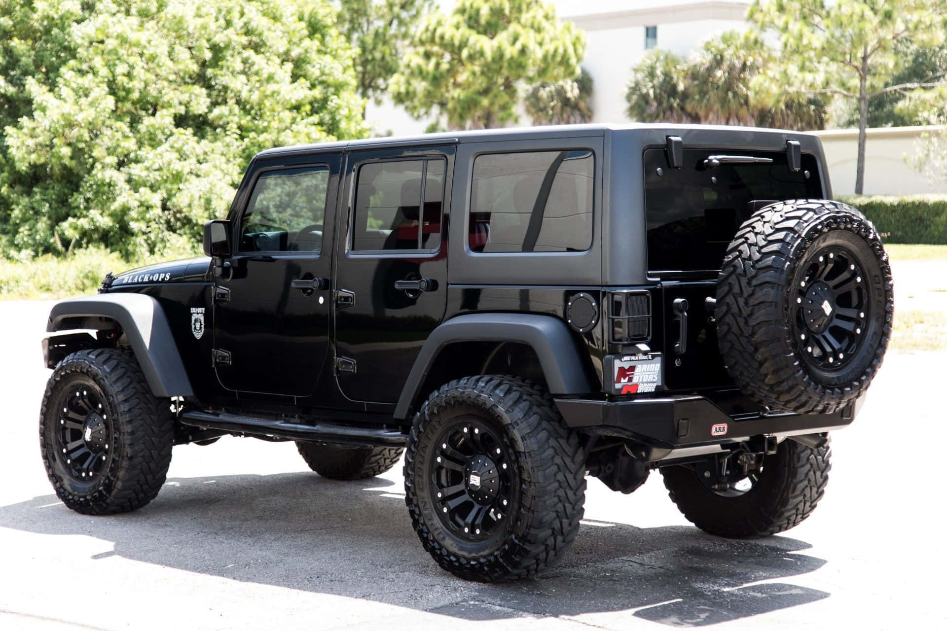 黑色吉普車 桌布 壁紙 — 一輛亮黑色升高底盤的Jeep Wrangler，配備超大輪胎，在明亮的日光下停在柏油路上，背景有綠樹。