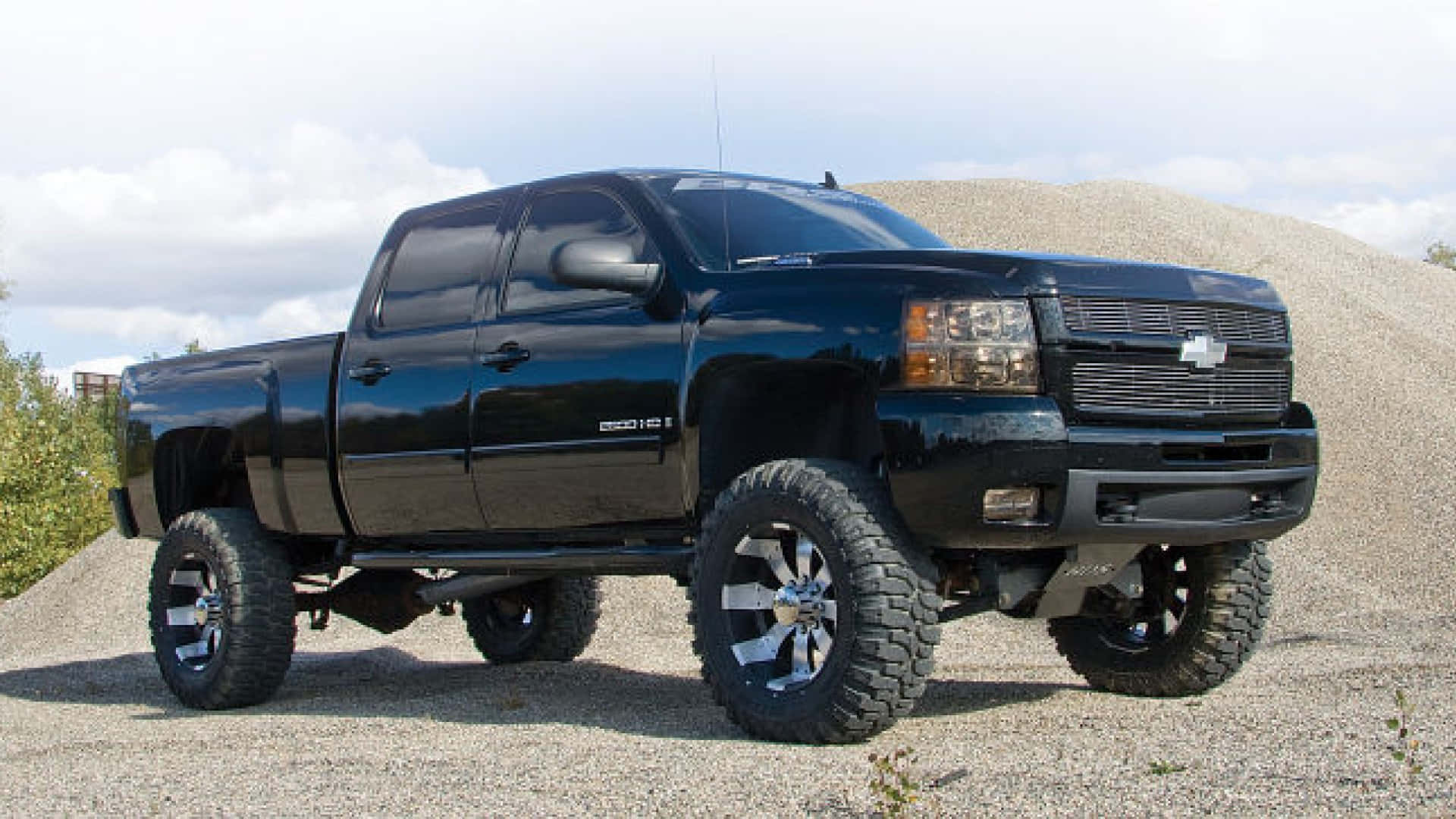 Jacked Up Trucks 桌布 壁紙 — 一輛亮黑色升高的Chevrolet Silverado皮卡車，配備大型越野輪胎，停在礫石堆附近的礫石地上，天空多雲。