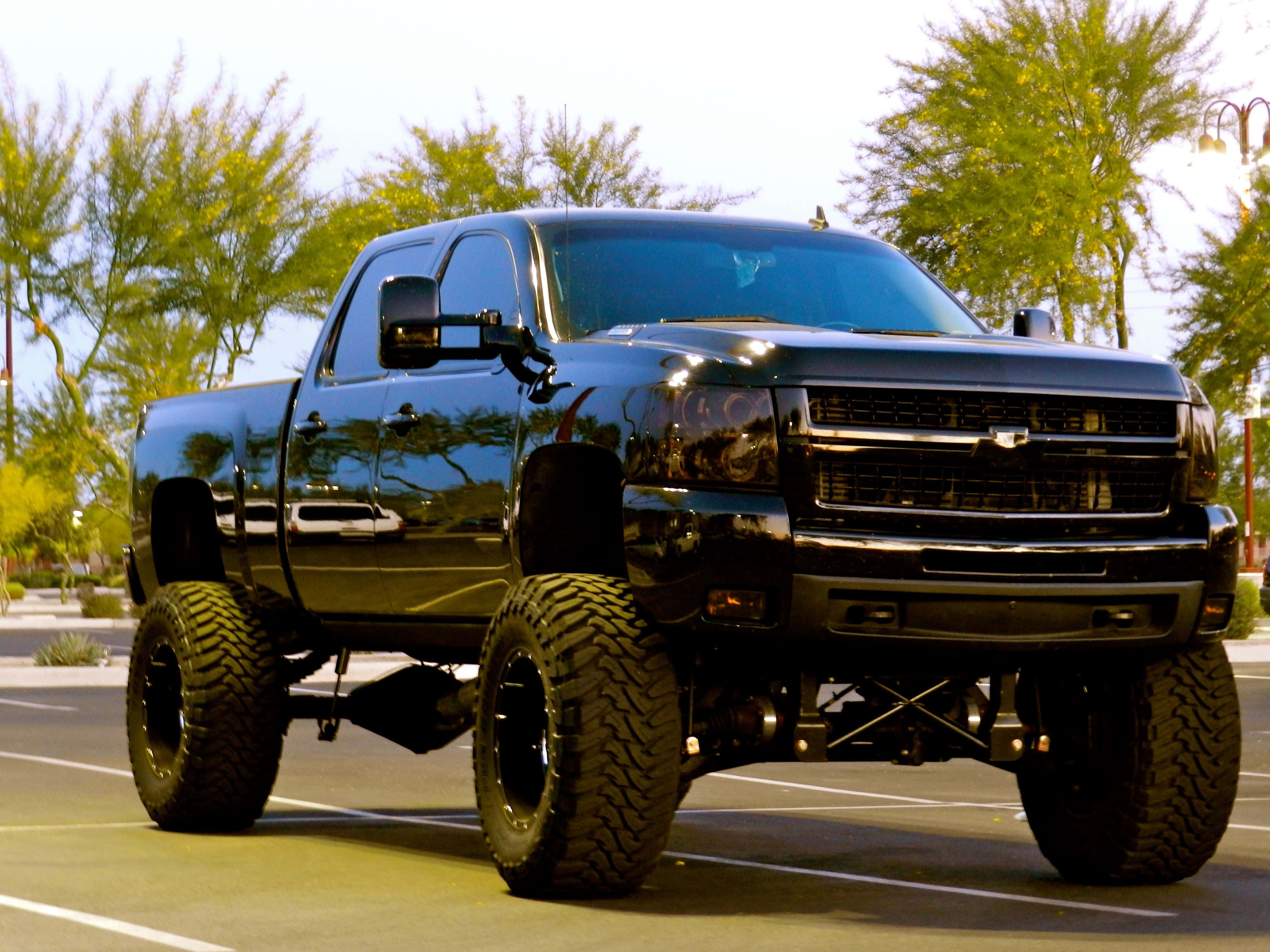 Lifted Truck 桌布 壁紙 — 一輛亮黑色升高的雪佛蘭皮卡車，配備巨大輪胎，停放在一條陽光普照、兩旁種滿綠樹的街道上。
