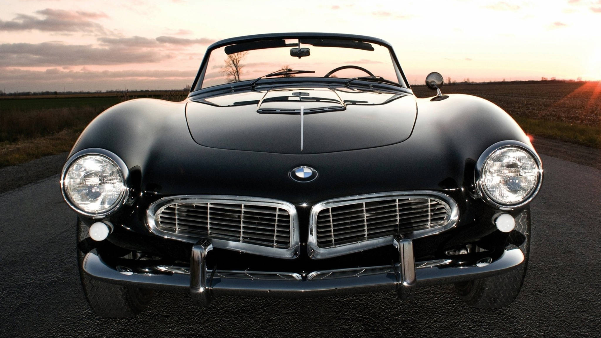 classic bmw 桌布 壁紙 — 一輛亮黑色復古寶馬敞篷車在日落時分於鄉村道路上向前行駛，天空色調溫暖。