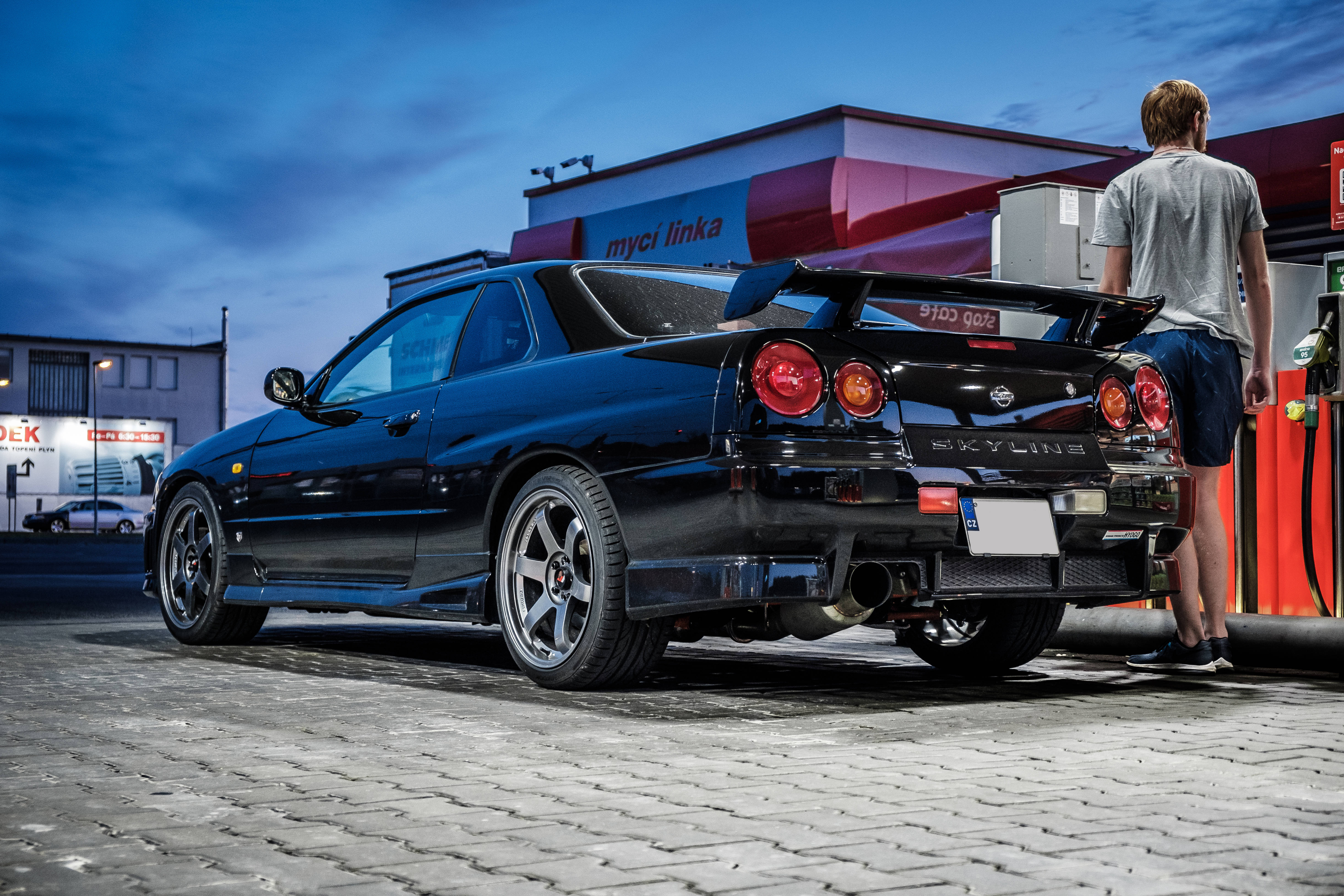 Black 桌布 壁紙 — 一輛亮黑色日產Skyline R34停放在加油站，一名男子在暮色下為其加油。