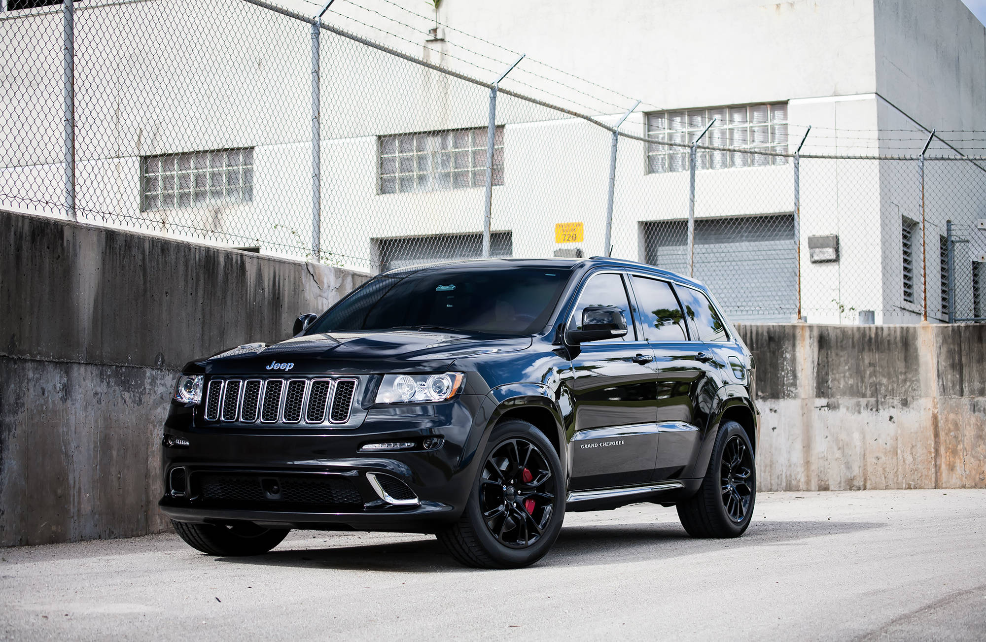 吉普 桌布 壁紙 — 一輛亮黑色的Jeep Grand Cherokee SRT停在鏈條圍欄和混凝土牆前，在明亮的日光下。