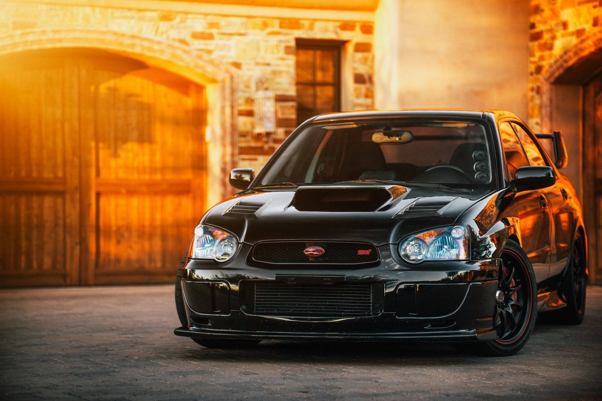 昴 桌布 壁紙 — 一輛亮黑色的Subaru WRX STI跑車在金色夕陽下，於質樸的石頭庭院中閃閃發光。