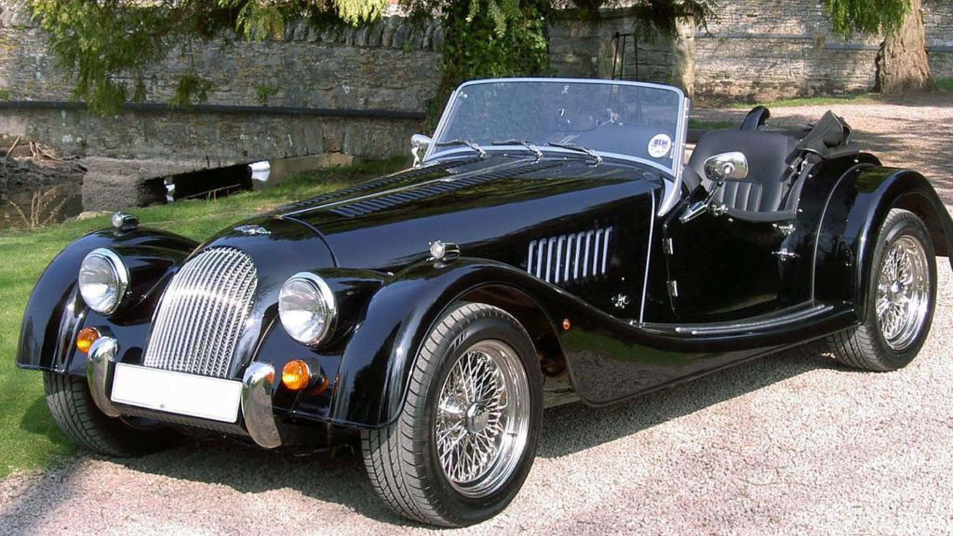 Morgan Roadster 桌布 壁紙 — 一輛亮黑色經典摩根敞篷跑車停放在碎石路上，旁邊有石牆和綠草。