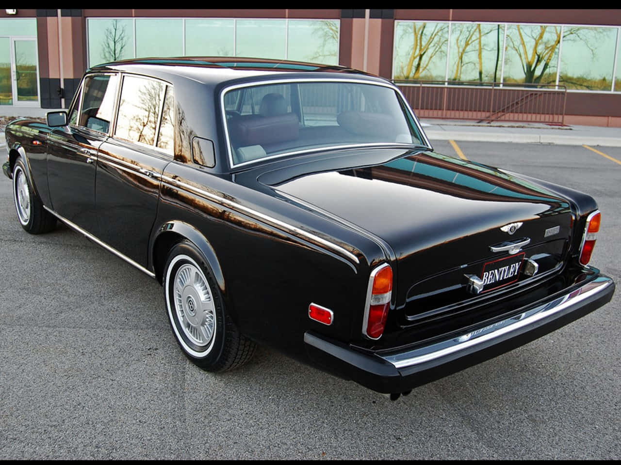 Bentley T2 桌布 壁紙 — 一輛亮黑色經典賓利轎車停放在現代化停車場，背景有反光窗戶和光禿禿的樹木。