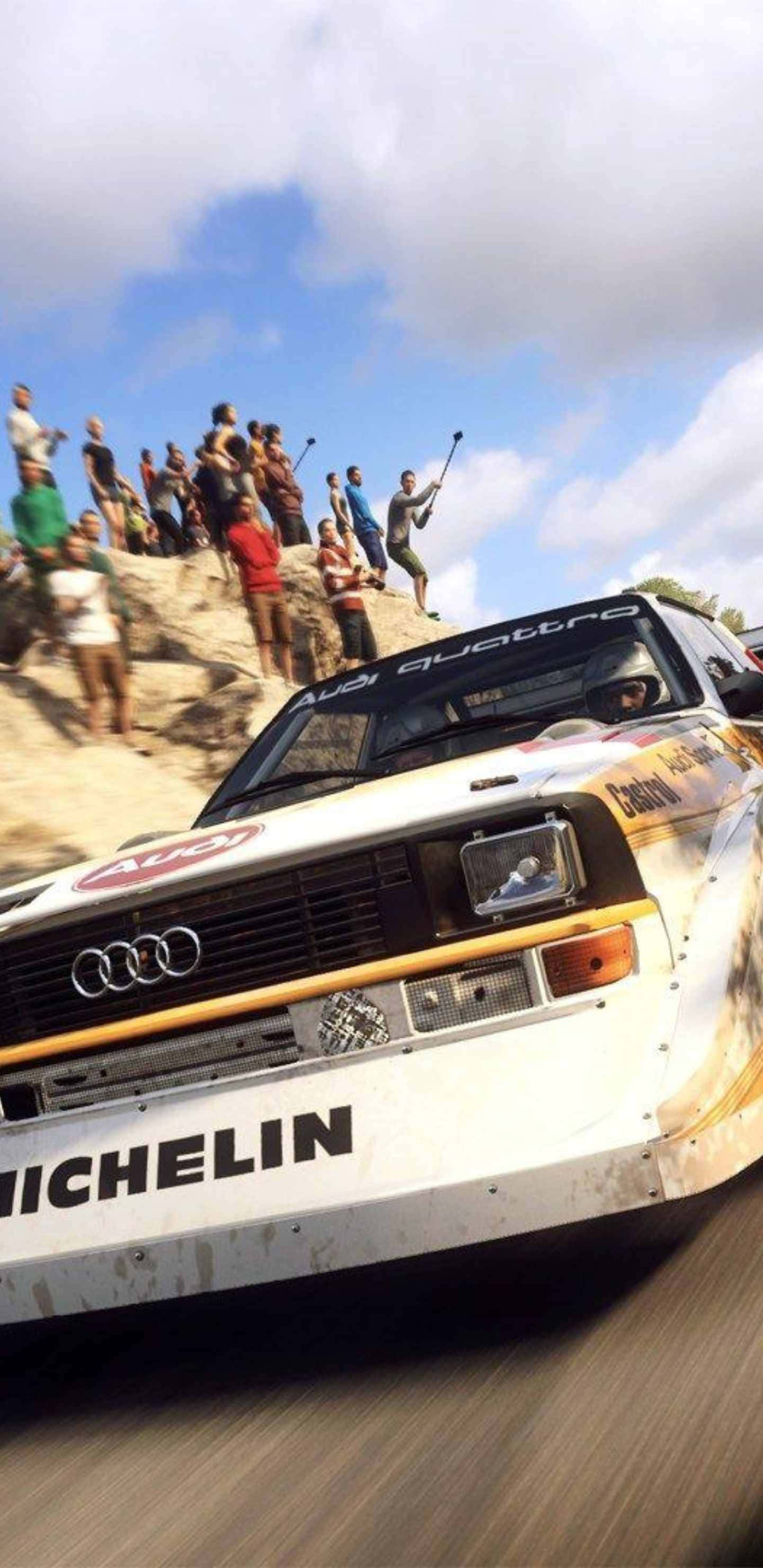 Android Dirt Rally 桌布 壁紙 — 一輛佈滿灰塵的白色奧迪 Quattro 拉力賽車，在明亮的藍天下，飛馳過坐在岩石山坡上的觀眾。