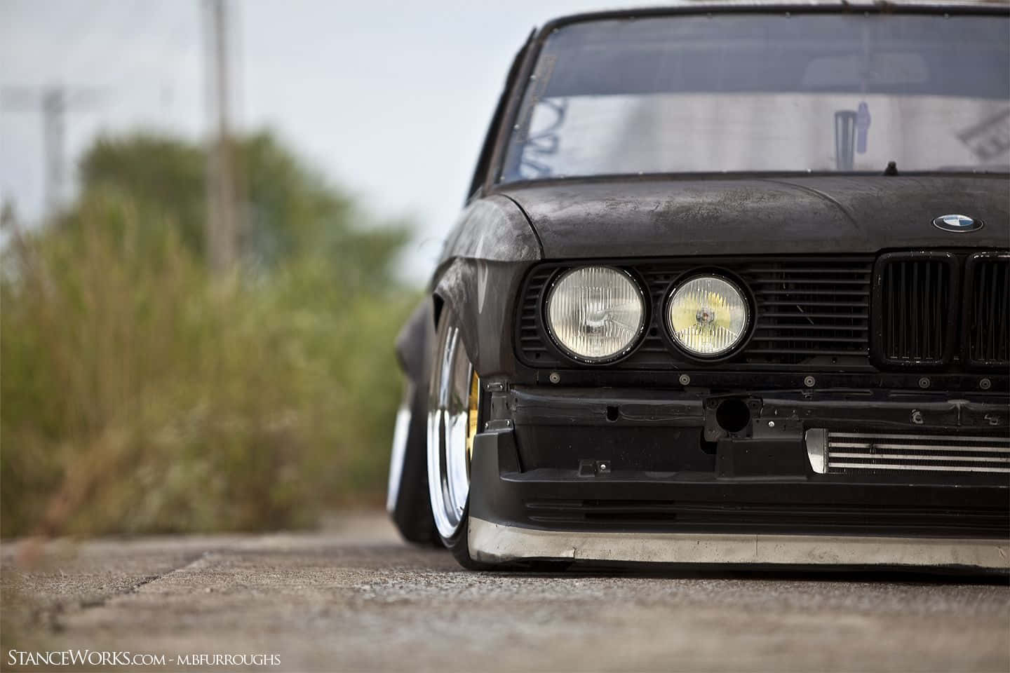 Stance Jdm 桌布 壁紙 — 一輛低矮的黑色BMW E30，配備訂製輪圈和風化塗裝，停在裂開的瀝青路上，旁邊是雜草叢生的草地。