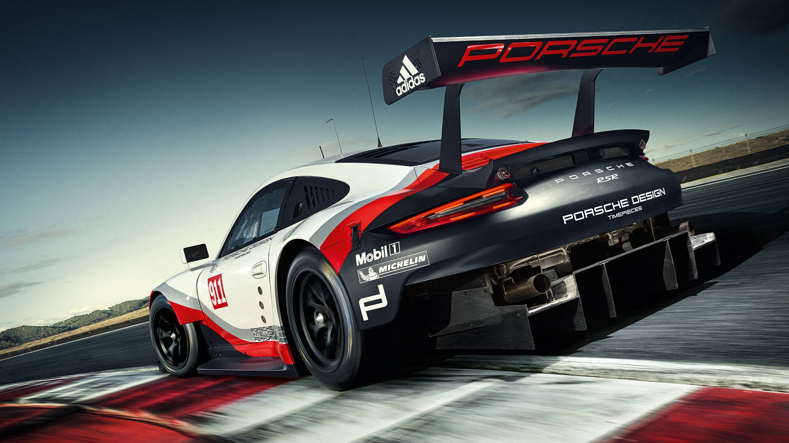 賽車 桌布 壁紙 — 一輛保時捷 911 GT3 R 賽車在晴朗的天空下沿著賽道飛馳，帶有動態模糊和紅白格子路緣。
