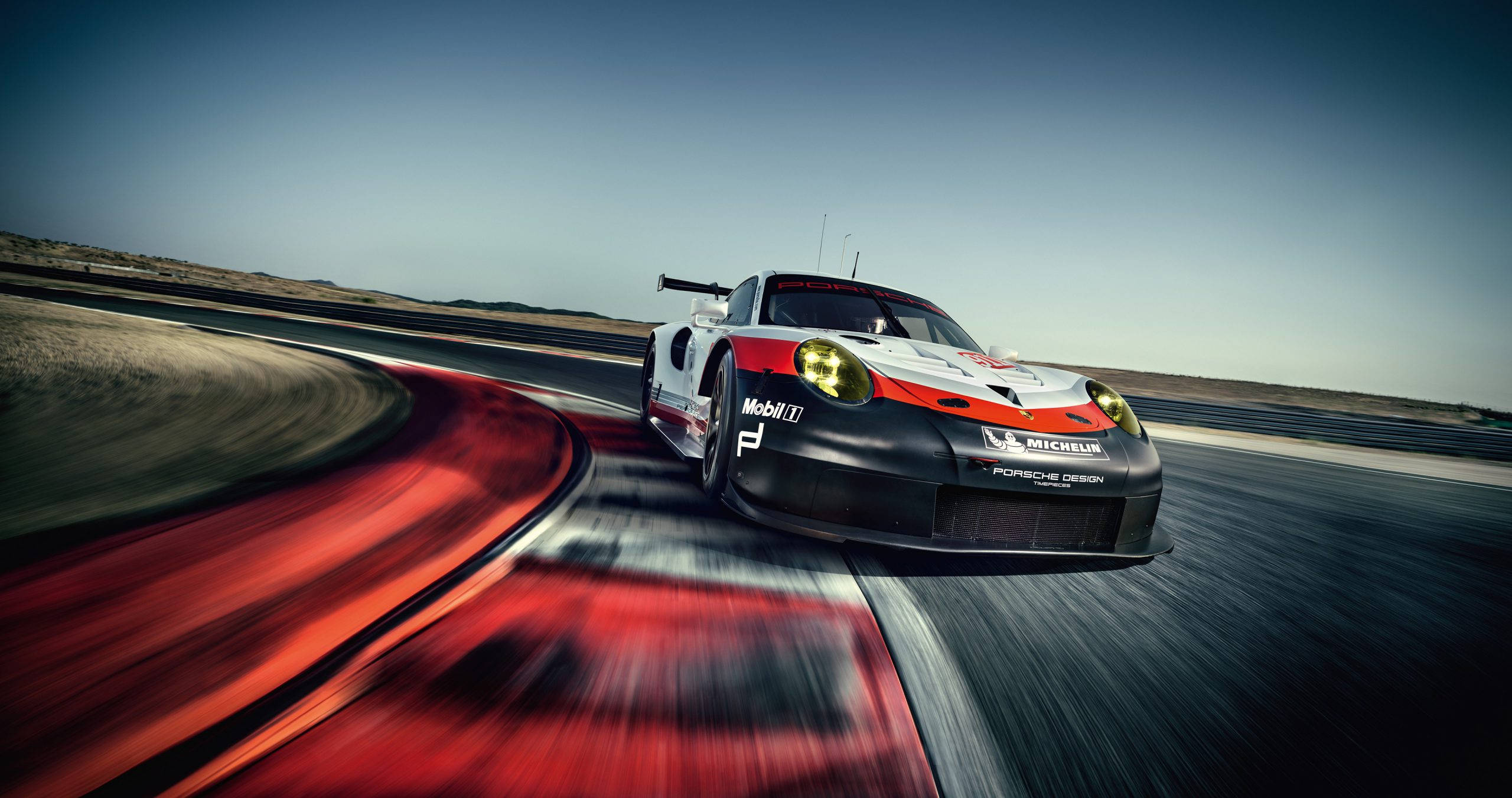 Auto Racing 桌布 壁紙 — 一輛保時捷賽車在沙漠賽道上疾馳，動態模糊的紅色和灰色在清澈的藍天下拖曳。