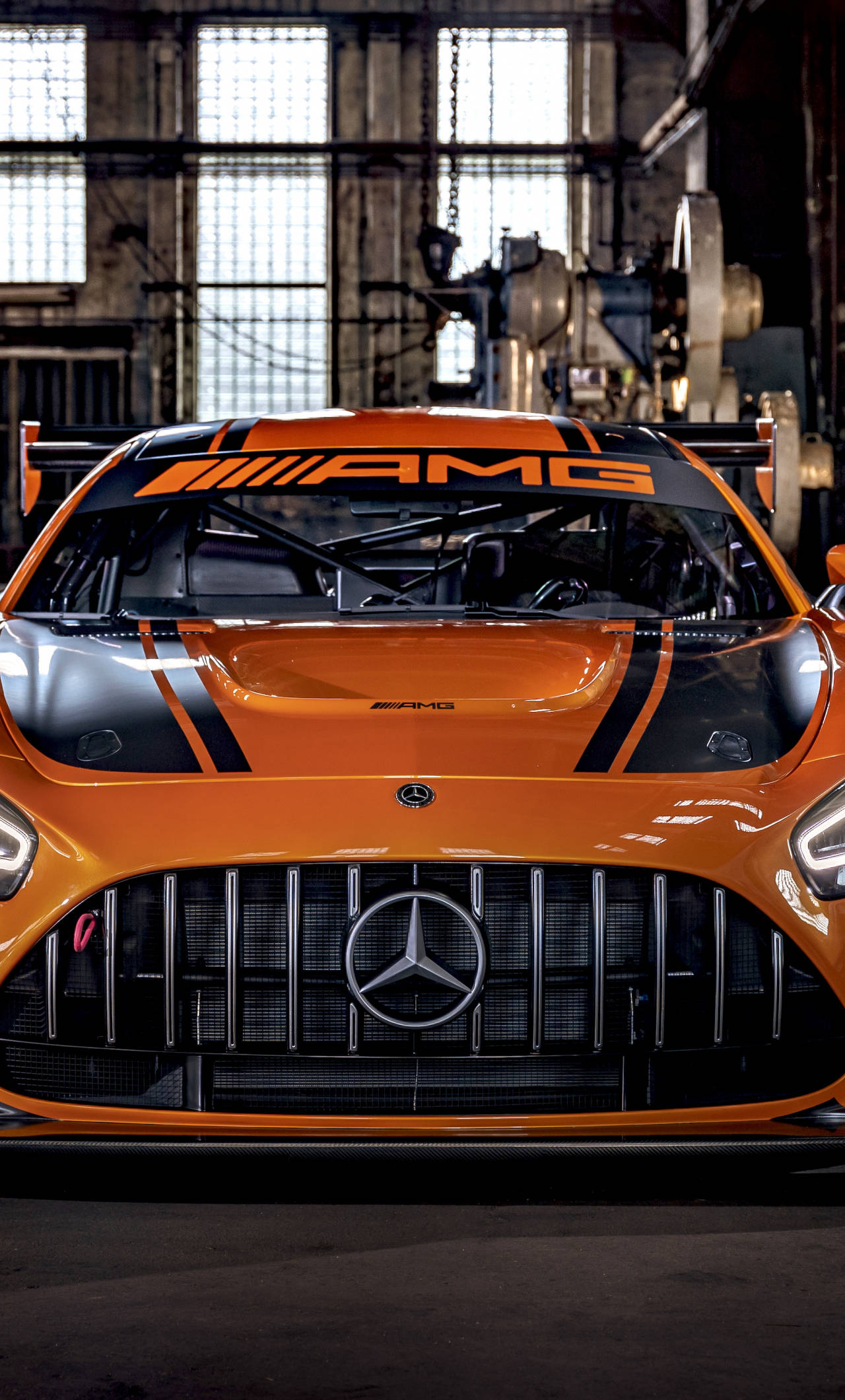 amg iPhone 桌布 壁紙 — 一輛充滿活力的橘色 Mercedes-AMG 賽車，帶有黑色點綴，在明亮的頭頂燈光下展示於骯髒的工業車庫中。