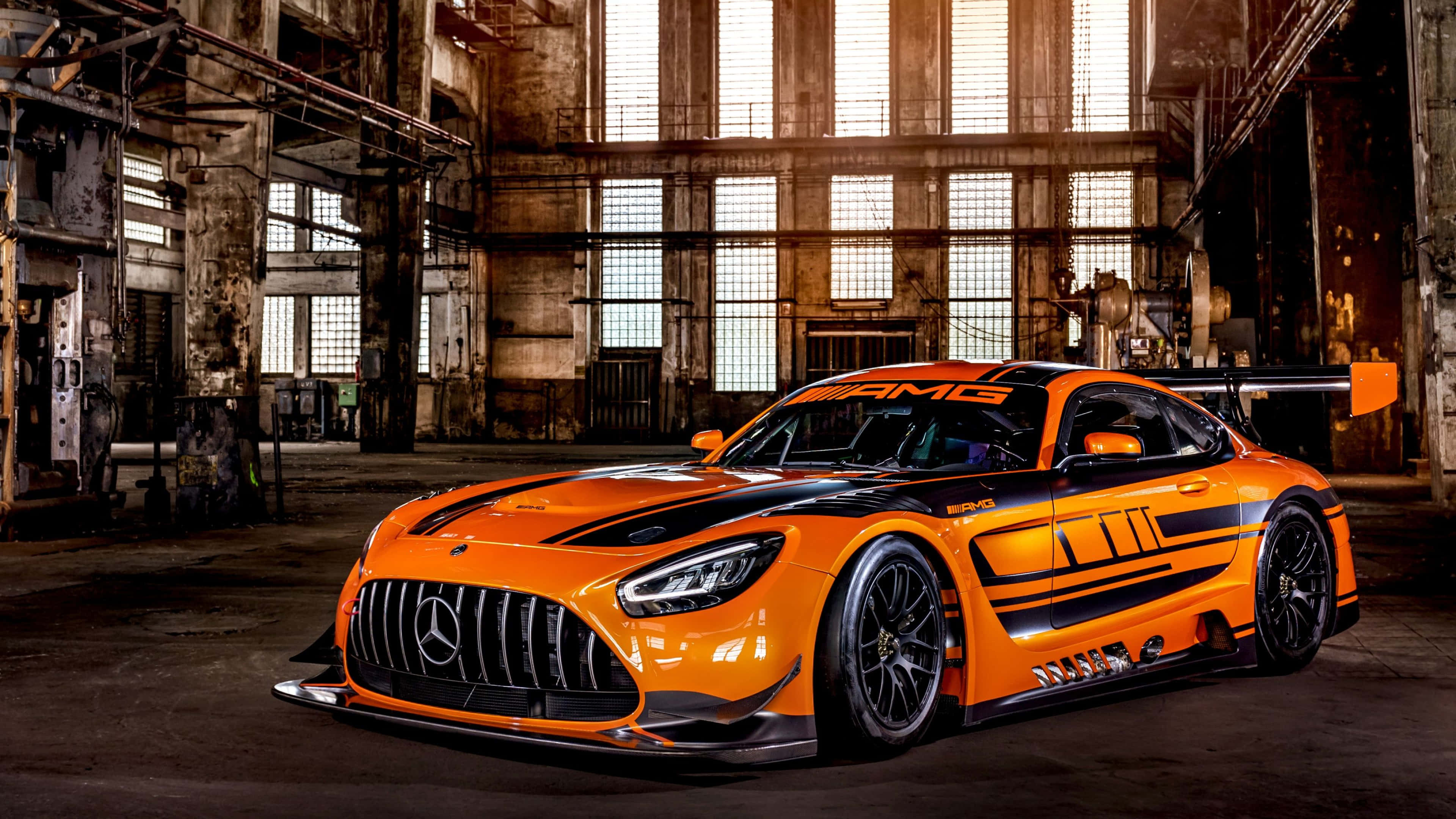 桌布 壁紙 — 一輛充滿活力的橘色 Mercedes-AMG GT3 賽車，帶有黑色點綴，戲劇性地停放在陽光普照的廢棄工業倉庫中。