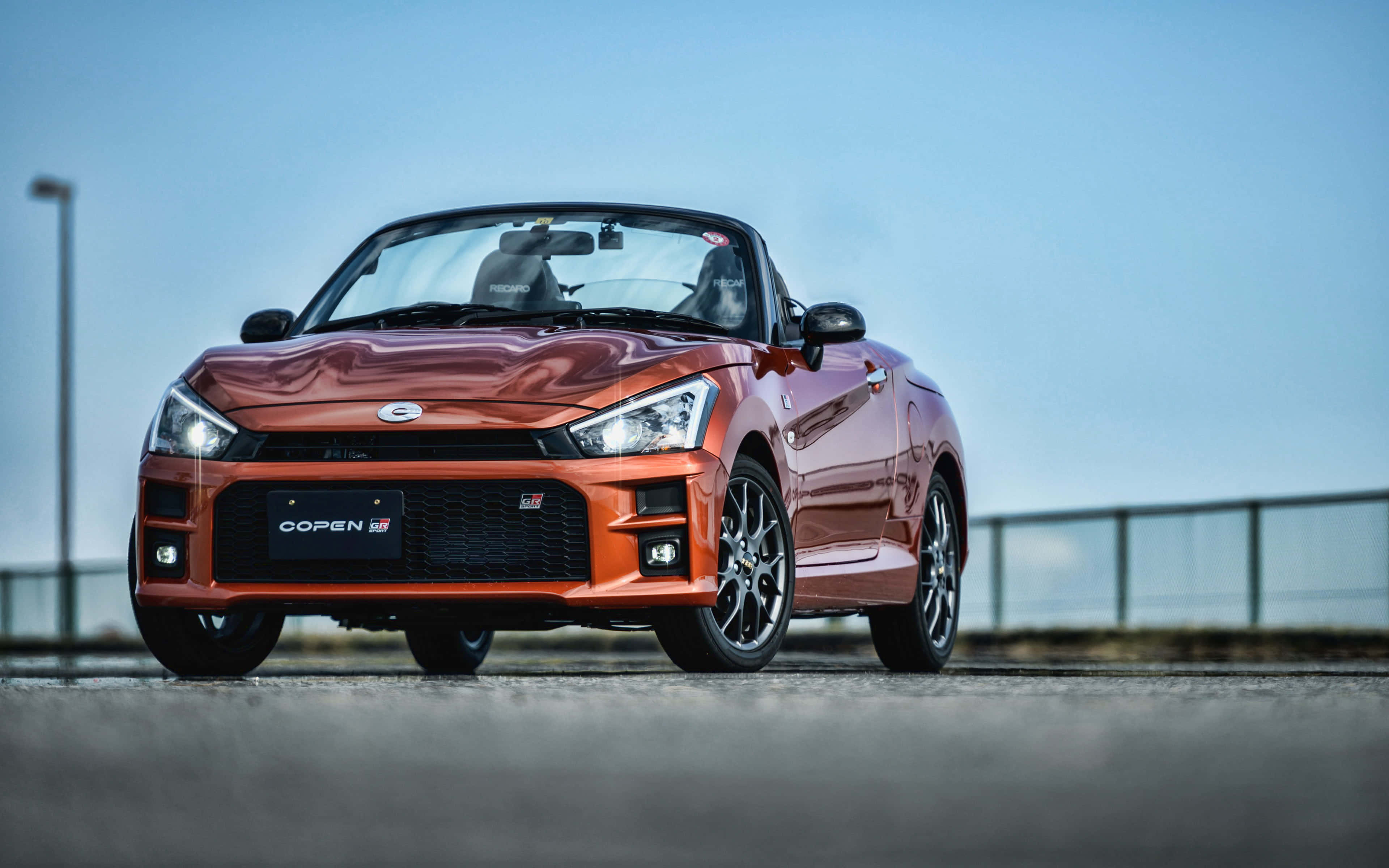 Daihatsu 桌布 壁紙 — 一輛充滿活力的橘色鈴木 Copen GT 敞篷跑車在晴朗藍天下閃耀於鋪砌道路上。